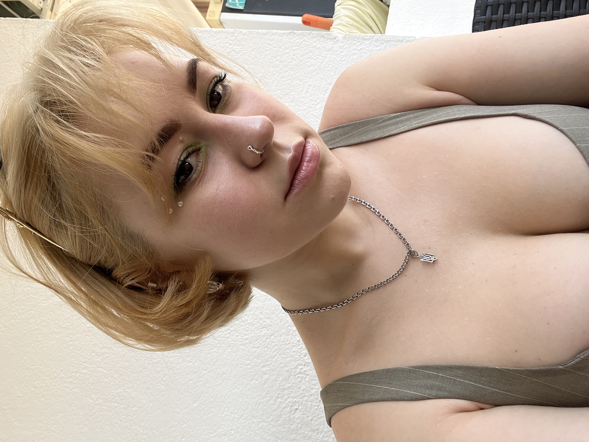 This close-up portrait features a young woman with blonde hair, styled with bangs, looking directly at the viewer. She has fair skin, dark eyebrows, and brown eyes. Her eye makeup is notable for its vibrant green eyeshadow, winged eyeliner, and delicate, clear gem-like embellishments positioned below her left eye. A silver hoop piercing is visible on her nose, and her lips are full with a soft pink hue.
She is wearing an olive green top with subtle vertical stripes, possibly a camisole or tank top. A silver chain necklace adorned with a small Hamsa hand pendant rests on her chest. A gold-toned hair accessory can be seen partially in her hair on the left side.
The woman's head is tilted, giving her a contemplative and direct gaze. The lighting is soft and natural, suggesting it is daytime. The background primarily consists of a plain white or light-colored wall, with faint hints of darker, possibly woven outdoor furniture or structures visible in the upper right, hinting at an outdoor or semi-outdoor setting. This could be a balcony or patio in Budva, Montenegro. There is no visible text in the image.