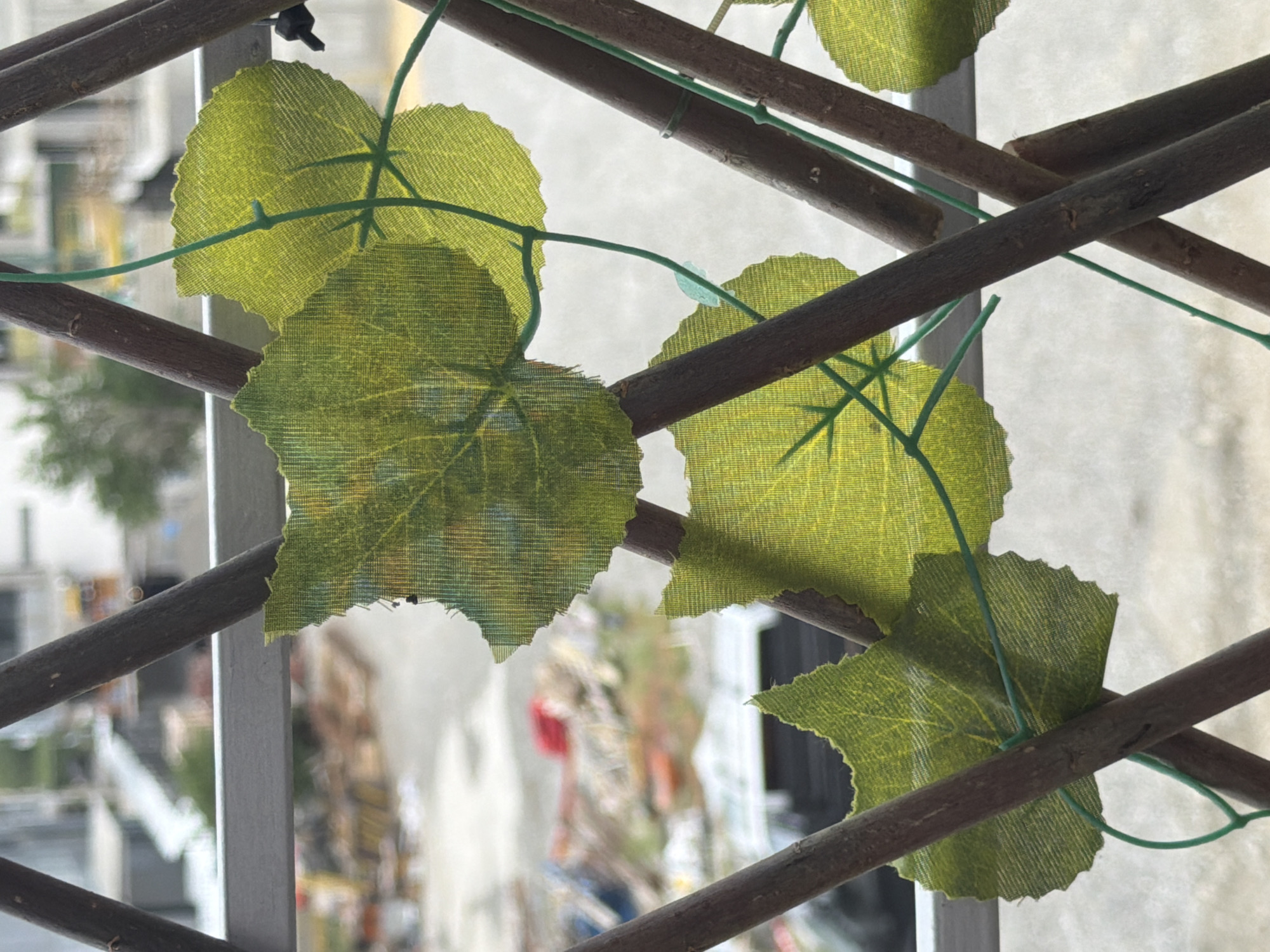 The image shows artificial green leaves attached to a wooden trellis structure. The trellis is made of dark brown, thin branches arranged in a criss-cross pattern. The leaves are a vibrant green with visible veins and are attached to the trellis with thin green wire. In the background, a blurred urban landscape is visible, suggesting an outdoor setting. The background includes buildings, construction materials, and trees, with an overcast sky. The scene appears to be on a balcony or a similar elevated outdoor space overlooking a city. It is not possible to determine the specific location in Tbilisi, Georgia, from the visual information provided.
