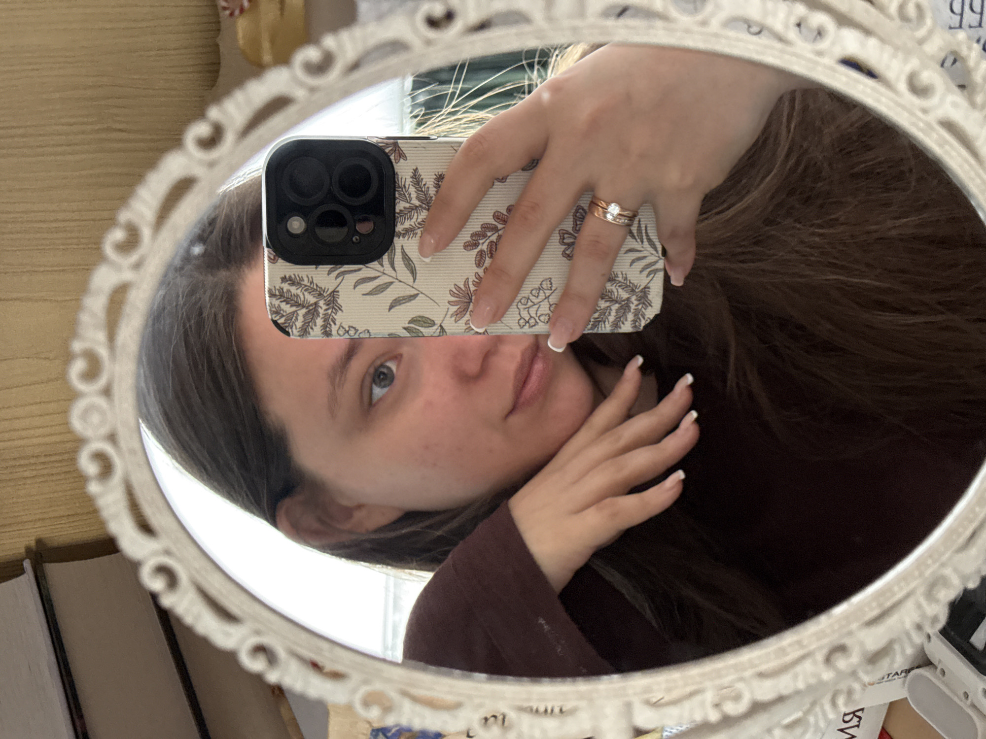 An overhead-angle mirror selfie depicts a female individual from the chest up, reflected in an ornate white oval mirror with a scrolled frame. The female has long dark brown hair and light skin, with her visible left eye being blue. She is wearing a dark reddish-brown long-sleeved top. Her left hand is raised, with fingers extended and palm facing her neck/chin area. Her right hand holds a smartphone, partially obscuring the right side of her face. The phone has a cream-colored case with a floral pattern featuring leaves and small flowers in shades of brown, pink, and green. The phone's camera module, featuring multiple lenses, is visible. On the ring finger of her right hand, she wears two rings: a plain rose gold band and a rose gold band set with a central clear stone flanked by smaller clear stones. Both hands exhibit manicured fingernails with white tips. The background reflected in the mirror includes stacked books on the upper left, a light-colored textured vertical surface resembling wood paneling or wallpaper on the upper right, and bright white light from an unidentifiable source, likely a window, appearing behind the subject's head. Partial, indistinct objects and Russian text are visible at the bottom and right edges of the mirror's reflection, including the partially visible letters "ИЯ" on the bottom left. The scene is located in an indoor residential setting in Krasnodar, Russia.