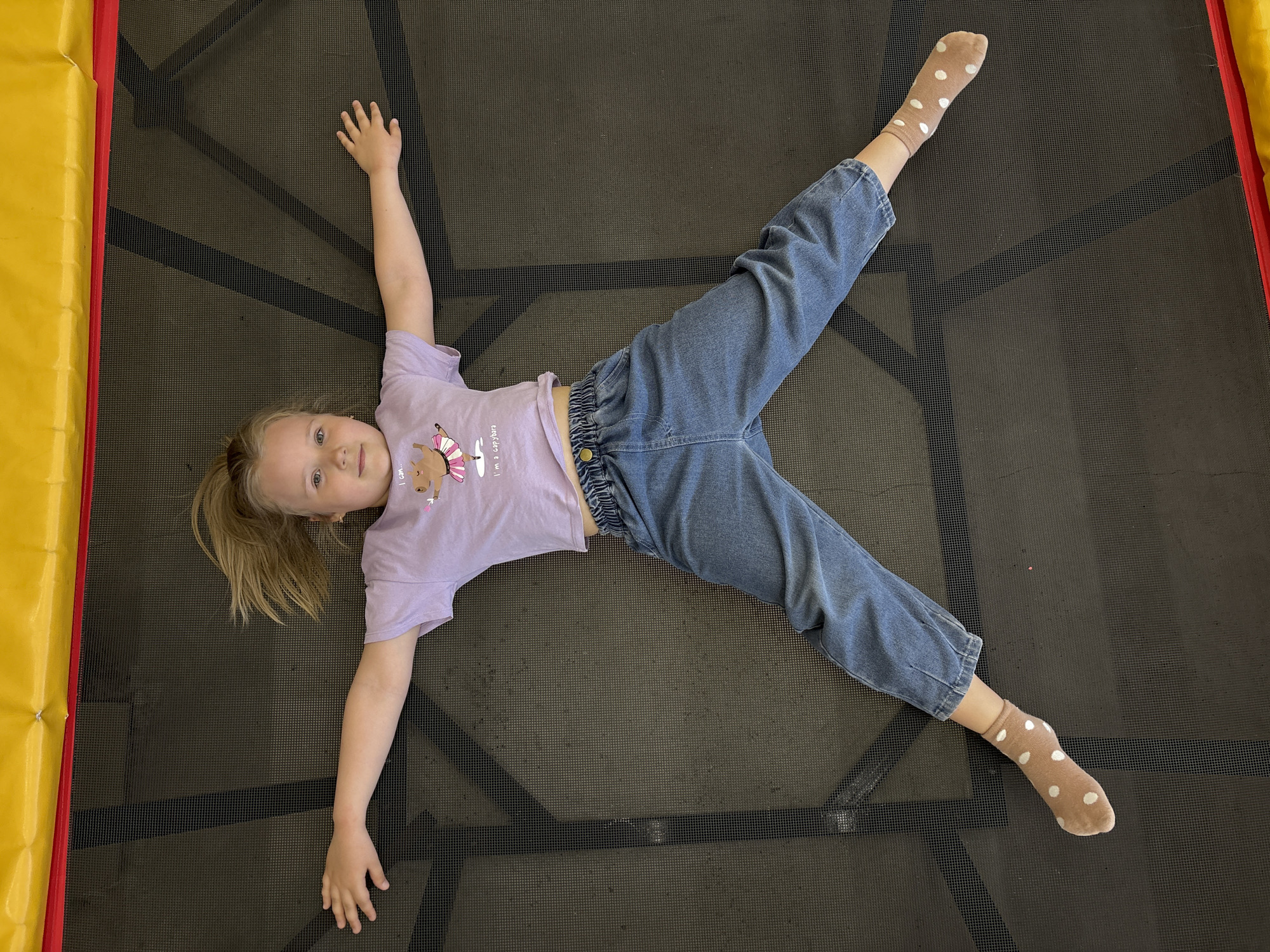 A young girl is lying in the center of a trampoline. She is wearing a light purple short-sleeved shirt and blue jeans, and her feet are clad in tan socks with white polka dots. The trampoline mat is dark gray. The frame of the trampoline is padded in yellow and red material.