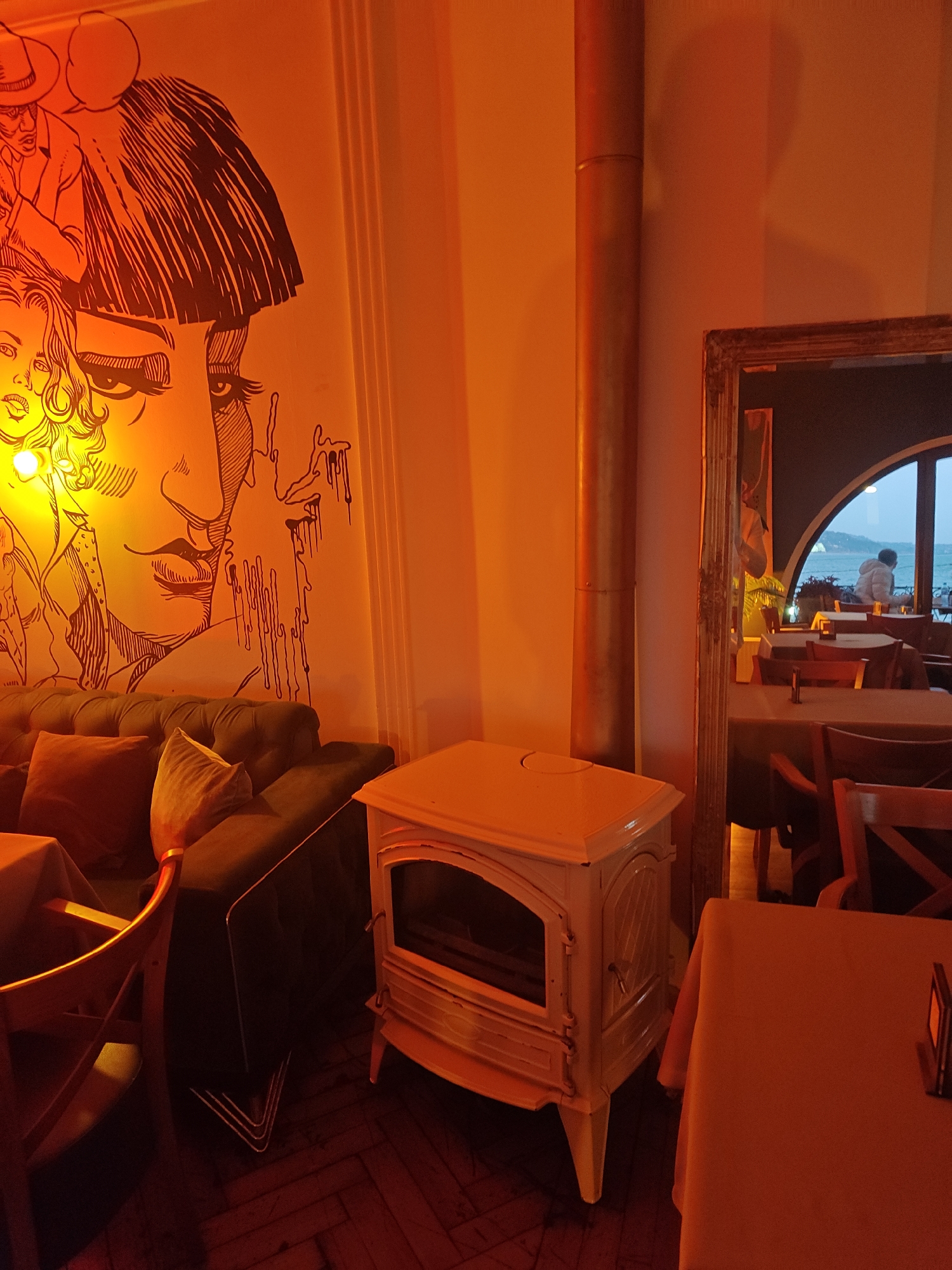The image captures a dimly lit, likely indoor, space with warm, possibly artificial, lighting that casts an orange hue across the scene. On the left, a large wall mural features a stylized, sketched portrait of individuals, with a sofa placed in front of the mural. The sofa is dark-colored, and is adorned with several pillows.
To the right of the sofa, a vintage-style white fireplace stands on the floor. A cylindrical duct runs vertically along the wall, suggesting a heating or ventilation system. A framed mirror reflects a view of the restaurant interior, including tables, chairs, and a window or doorway in the background where a person is visible near what appears to be a body of water or the sea.
The floor appears to be tiled or wooden. A table with an orange tablecloth and a wooden chair are visible in the foreground. The location is Unknown, Unknown.
