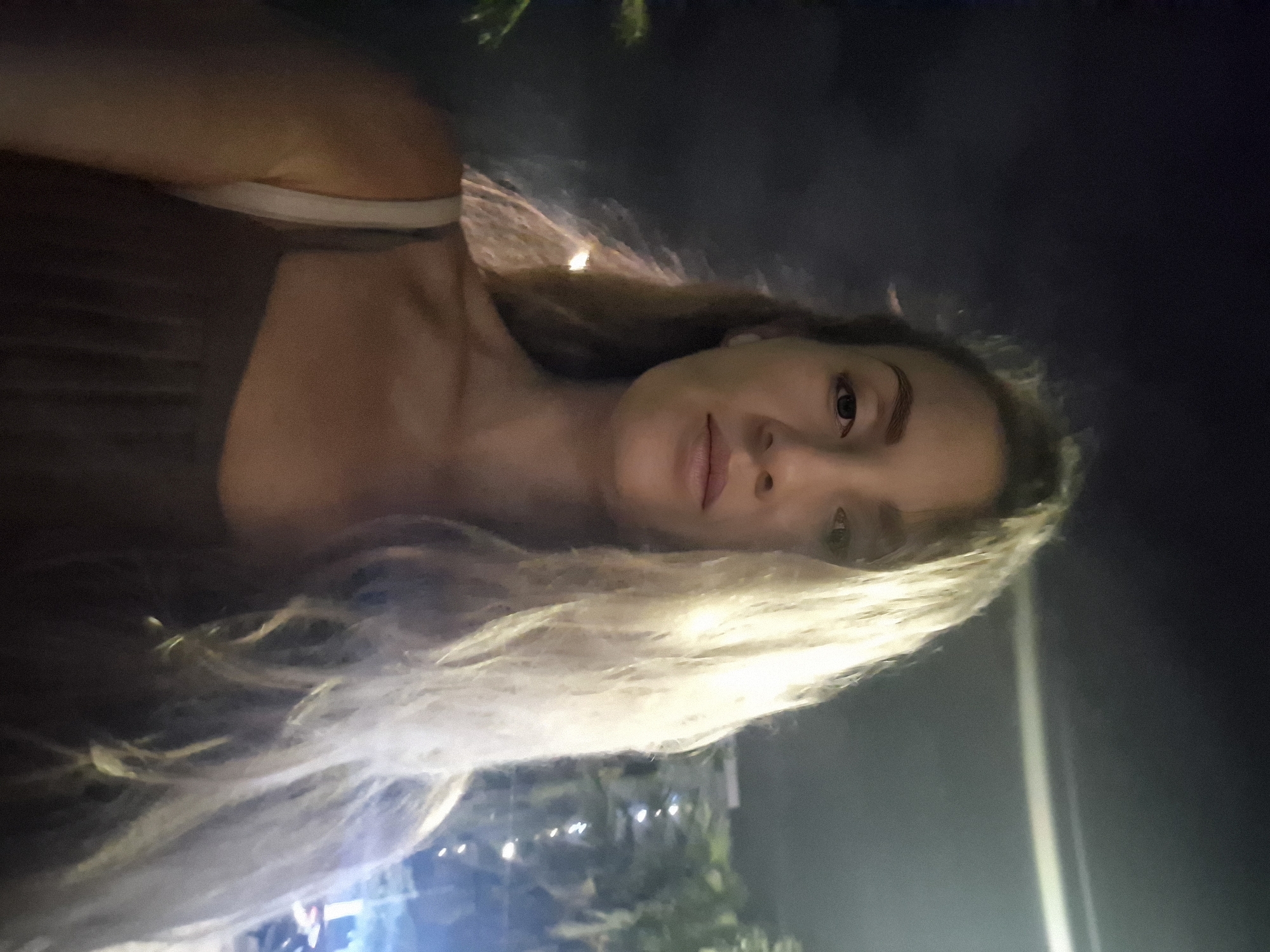 A selfie of a woman with blonde hair is taken at night in the city of Đà Nẵng, Vietnam. The woman is facing the camera, with her head slightly tilted. Her hair is long and wavy, and it falls over her shoulders. She is wearing a dark-colored top. In the background, there are palm trees and buildings illuminated by lights. The sky is dark and cloudy.