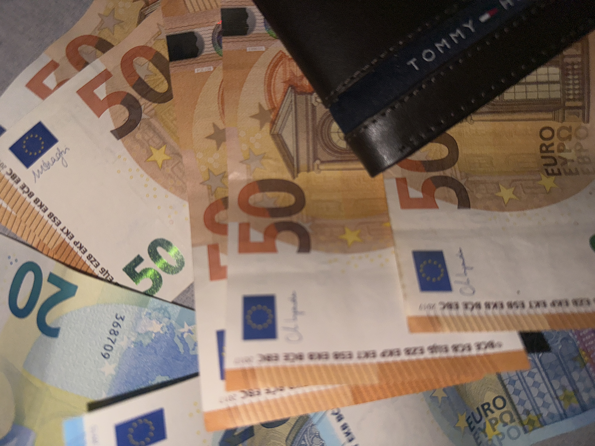The image shows a collection of Euro banknotes, specifically 50 Euro and 20 Euro notes, partially covered by a black Tommy Hilfiger wallet. The banknotes are scattered and slightly overlapping. The image is taken at a close-up angle, focusing on the money and the wallet. The background is indistinct.