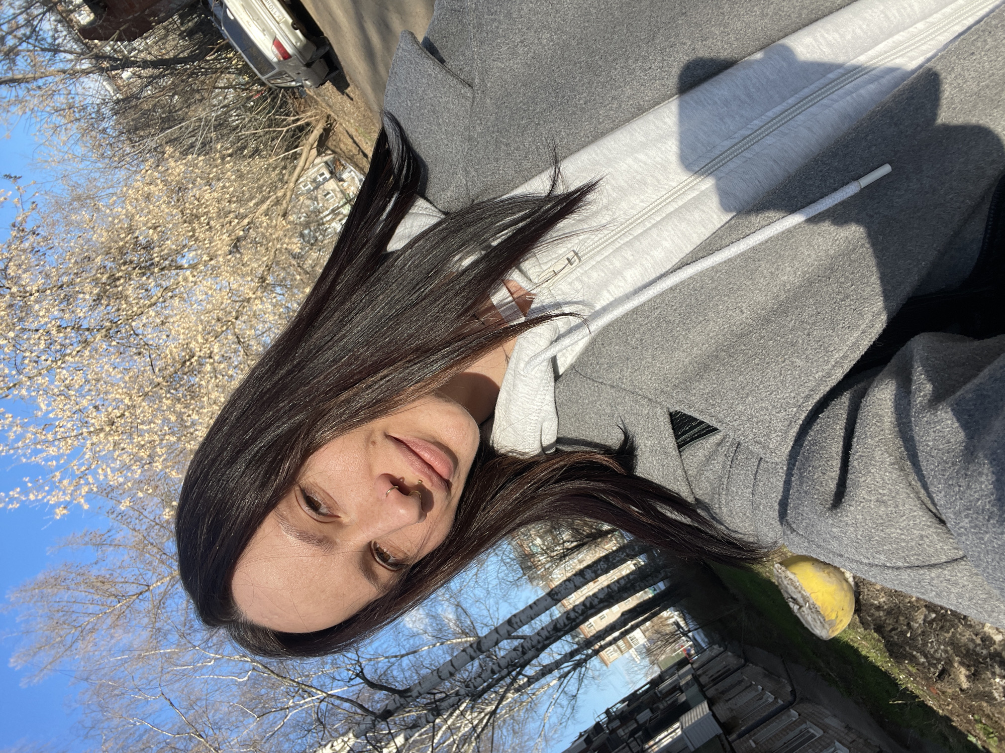 A woman with long dark hair, wearing a gray hooded jacket, is looking at the camera. She has a nose piercing. The background shows a street scene, with trees, buildings, and a car parked. The photo was taken in Izhevsk, Russia.