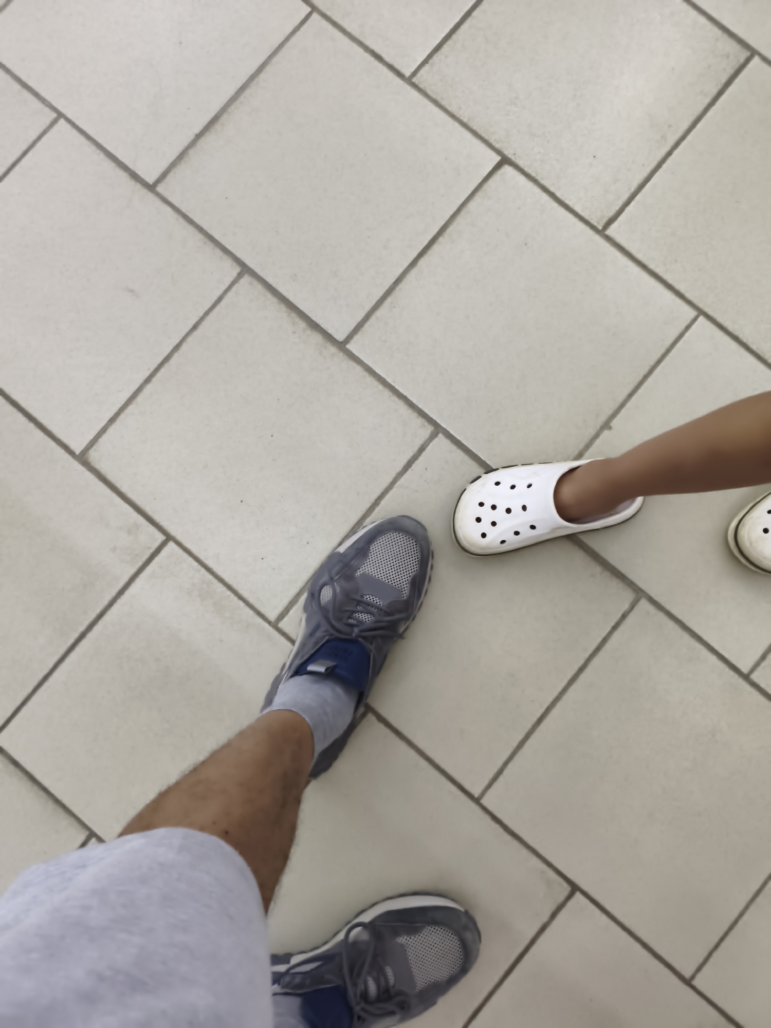 The image shows a first-person perspective of two individuals standing on a tiled floor, likely indoors. One individual is wearing grey athletic shoes and light-colored shorts, with their legs and feet visible. The other individual, presumably a child based on the size of their footwear, is wearing white Crocs. The floor is covered in large, square, light-colored tiles with dark grout lines arranged in a staggered pattern. The scene appears to be in the city of Aktobe, Kazakhstan, possibly in a public space or a home.