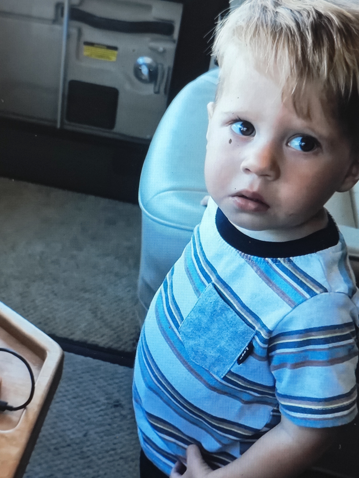 A child is photographed from the chest up, wearing a blue and white striped t-shirt with a black collar. The child has light brown hair and is looking slightly to the right with a pensive expression. A dark mole is visible on the child's left cheek. In the background, a light-colored seat and a piece of machinery with a yellow sticker are partially visible. The setting appears to be indoors, possibly in a vehicle or a room with carpeted flooring. There is no discernible indication of the location being Yarmolyntsi, Ukraine.