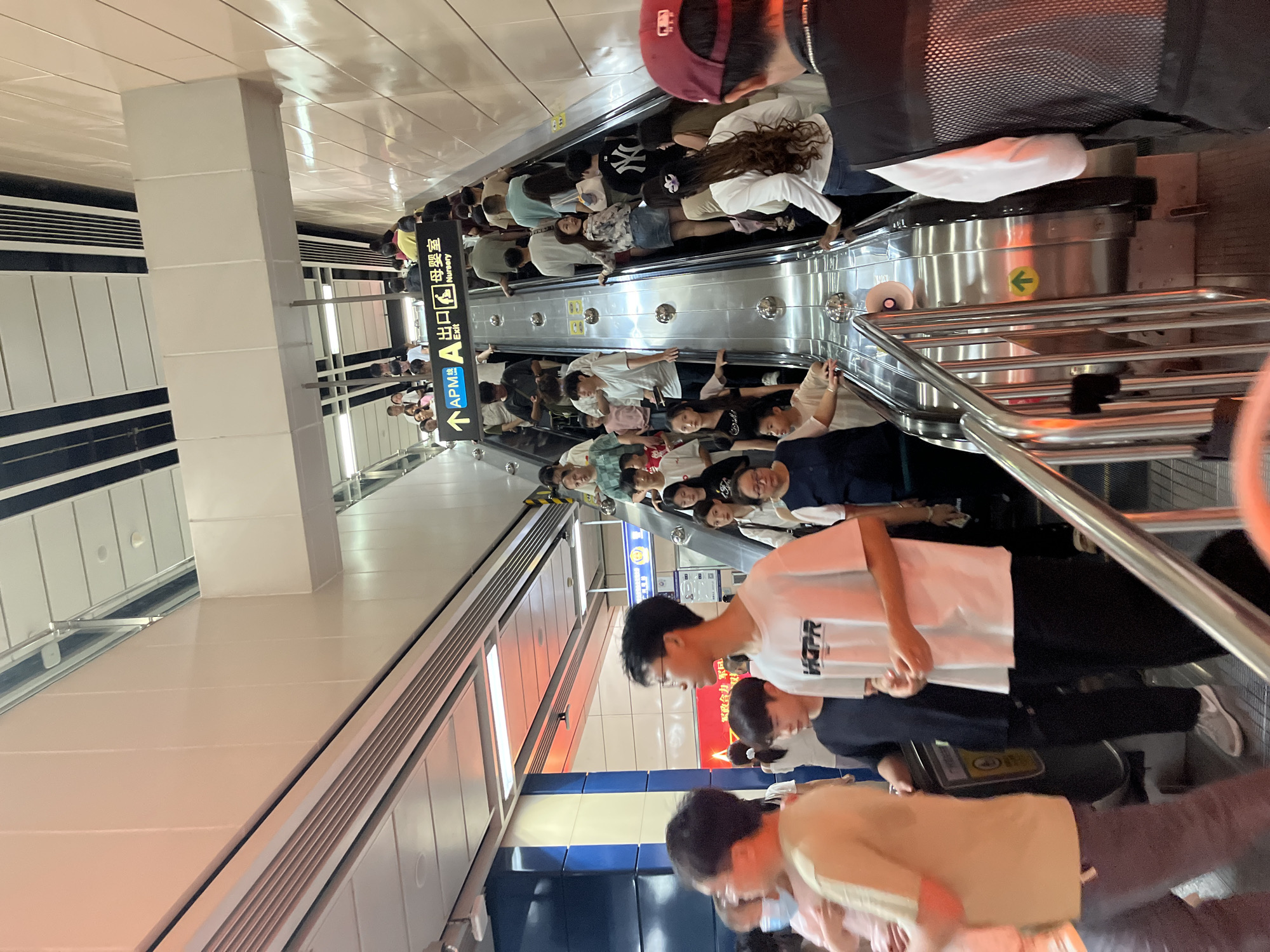 This image depicts a busy escalator in a subway station, likely in Haizhu District, China. A diverse group of people, of various ages and ethnicities, are ascending the escalator. They are dressed in casual, modern clothing. Some individuals are looking at their phones, while others are conversing with each other. The escalator is made of polished metal and has silver handrails. Above the escalator, a sign indicates "APM A Exit Nursery" in Chinese and English, suggesting it's a transportation hub with facilities for parents and children. The station environment appears clean and well-lit, with bright overhead lighting and pale walls, contributing to a modern aesthetic. The overall scene conveys a sense of urban transit and the daily flow of city life.