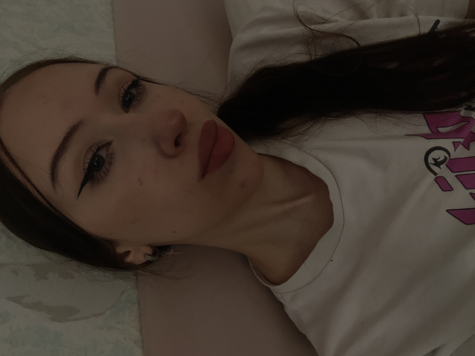 A young woman with long, dark hair is shown in a close-up, self-taken photograph. She is wearing a white t-shirt with a pink and black graphic design. Her makeup includes winged eyeliner and lipstick. She has a silver hoop earring with a cross charm in her left ear. The background appears to be a wall with a textured pattern and a pink surface, possibly a bed or couch. There is no discernible information about the location in Votkinsk, Russia, within the image itself.