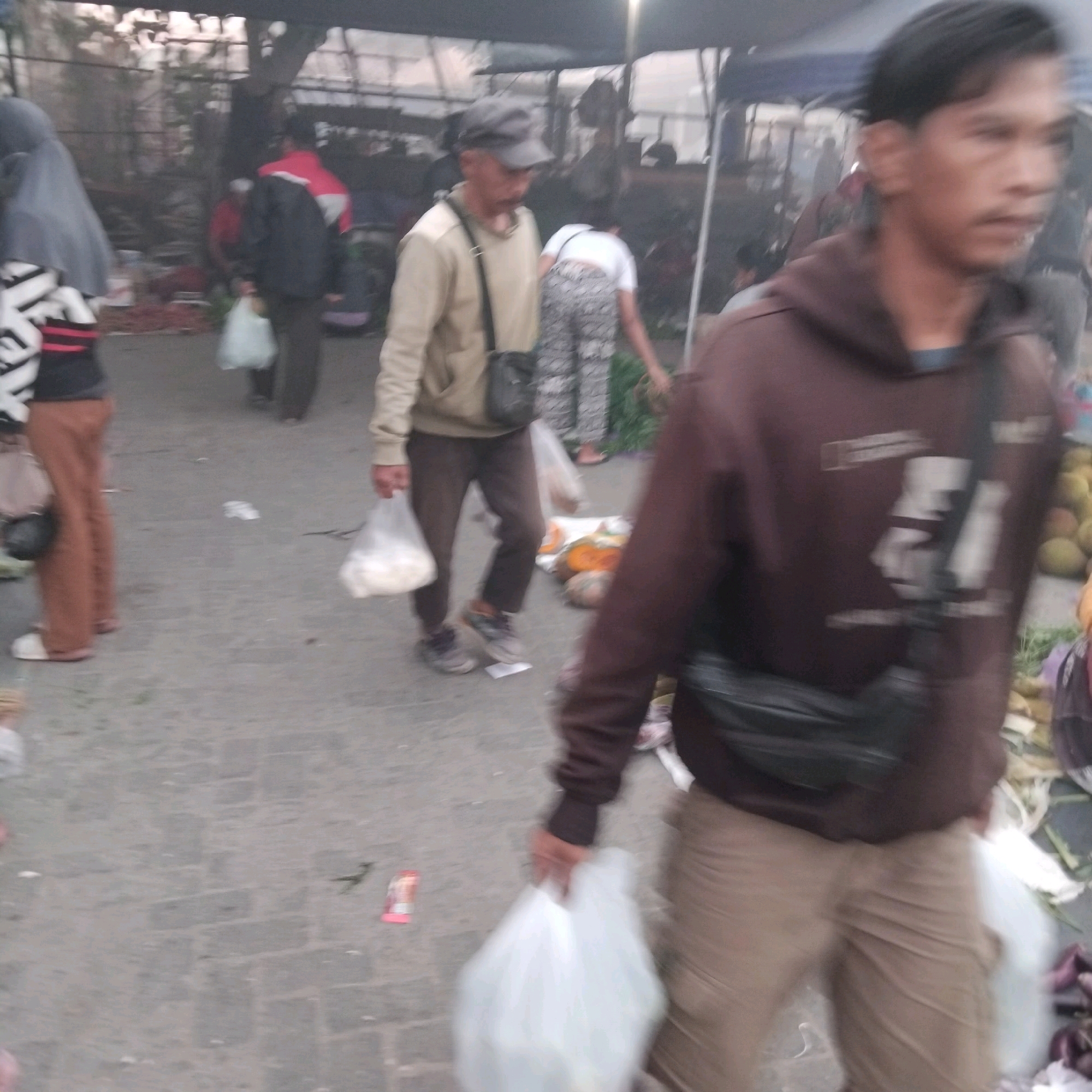 A bustling outdoor market in Banjarbaru, Indonesia, is depicted. Several individuals are visible, some carrying white plastic bags, suggesting they are either shopping or selling goods. One man in the foreground wears a brown hooded sweatshirt and light-colored pants, with a dark bag slung across his chest. Behind him, another man in a light-colored jacket and dark pants is also holding a white bag. To the left, a person in a grey headscarf and a patterned top stands with brown pants. The ground is paved with concrete blocks, and various items, including what appear to be fruits or vegetables, are scattered around, indicative of a marketplace. Tents or awnings are present in the background, providing shade.