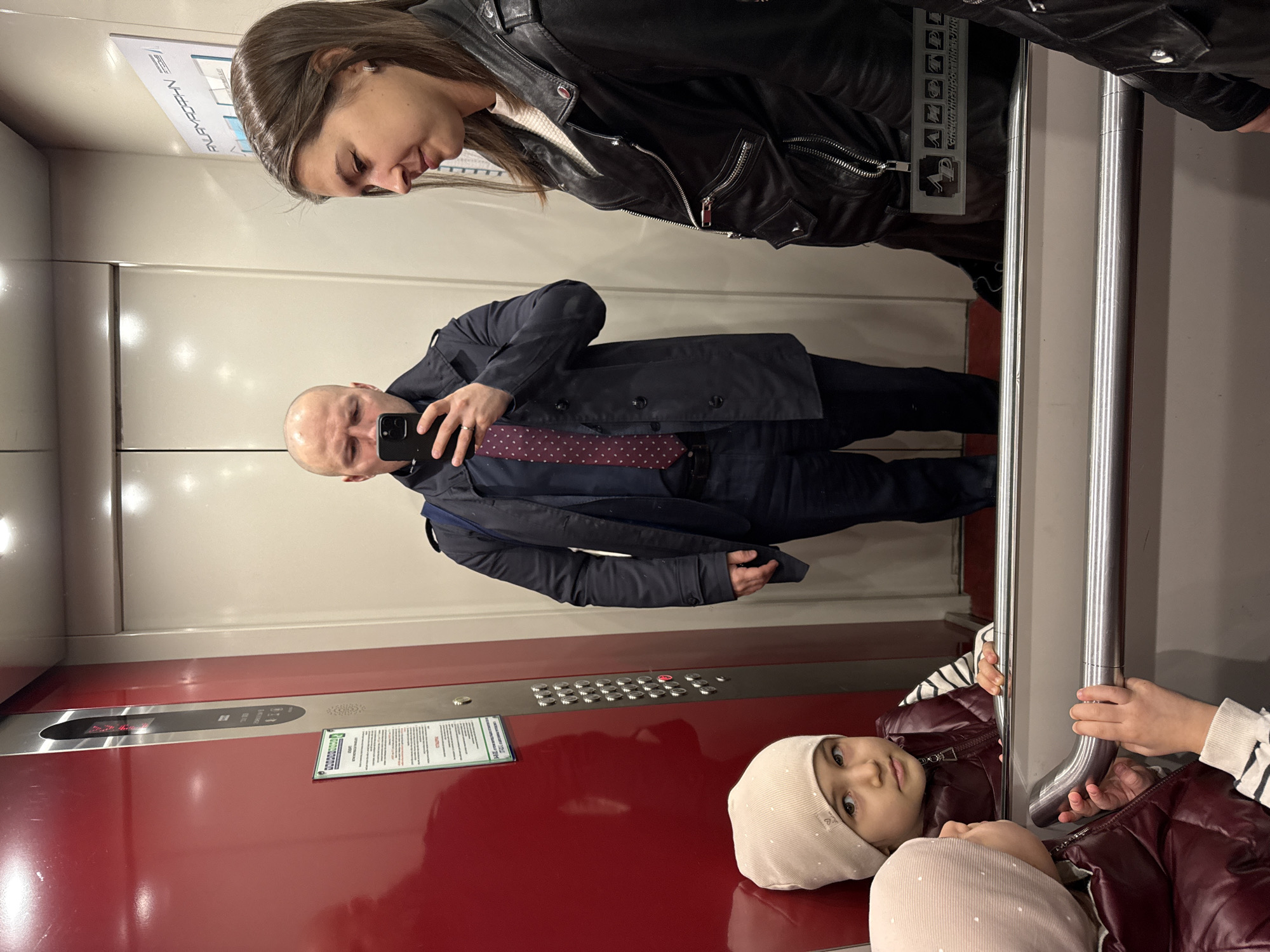 The image shows a reflection inside an elevator. A bald man in a suit and tie holds a phone taking a picture. A woman in a leather jacket is above him. A young girl with a hat and burgundy jacket is in front. The location is Krasnoyarsk, Russia.