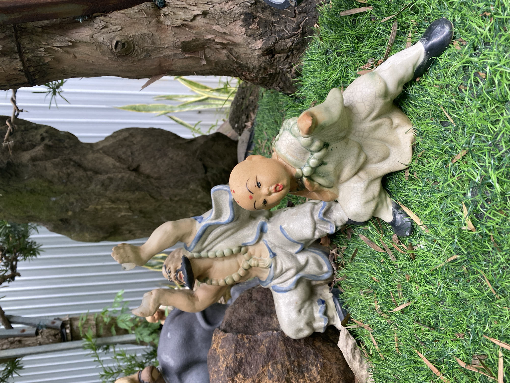 A statue of a bald man with a black beard and a string of beads around his neck is kneeling on artificial grass. He is wearing a white robe with blue trim and has his arms raised in the air. To his right, a smaller statue of a bald child with a red dot on its forehead is also kneeling on the grass. The child is wearing a light green robe and has its arms outstretched. In the background, there is a large tree trunk and a corrugated metal fence. A large brown rock is positioned to the left of the statue of the man. This scene is located in the city of Đà Nẵng, Vietnam.