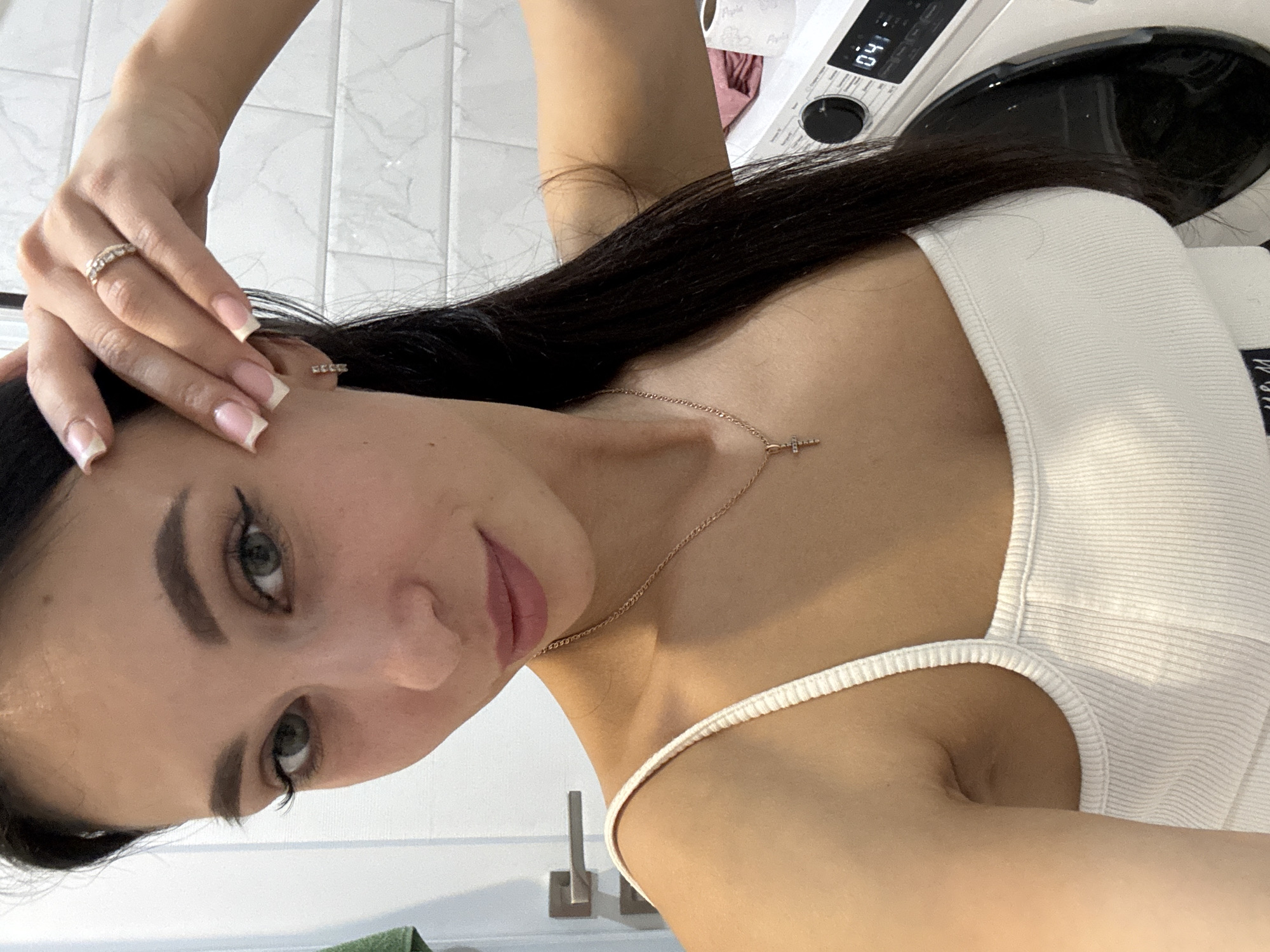 A woman is taking a selfie in a bathroom. The background shows white tile walls, a washing machine, a sink faucet, and a green towel. The woman is wearing a white tank top and a necklace with a small cross. She has dark hair, green eyes, and pink lipstick. She is wearing rings on her fingers and earrings. The time displayed on the washing machine is 04:11. There is no evidence indicating a specific location in Chaikovsky, Russia.