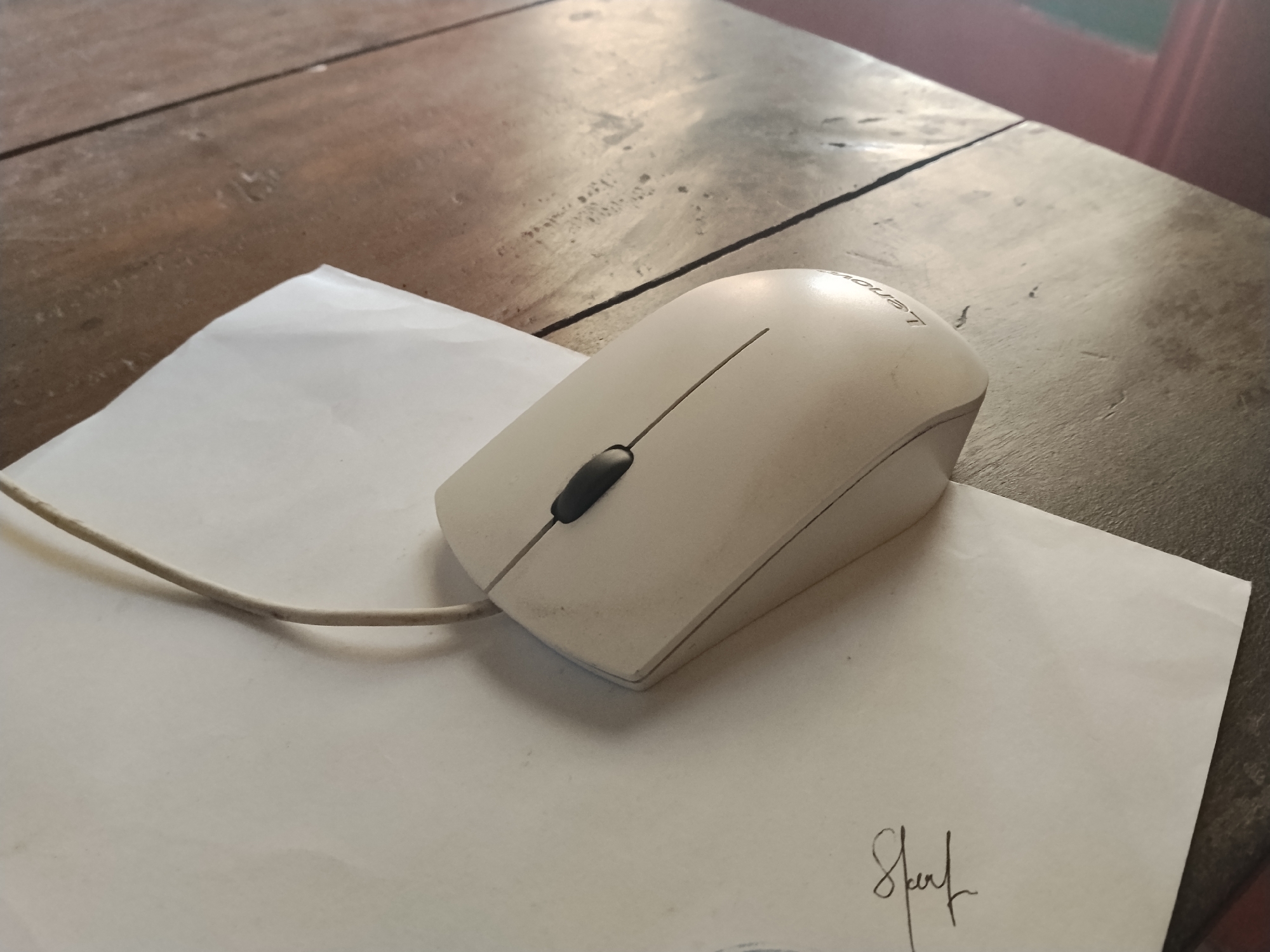 A white computer mouse with a black scroll wheel sits on a sheet of white paper. The paper is on a dark wooden table. A faint signature is visible on the bottom right of the paper. The scene is indoors.