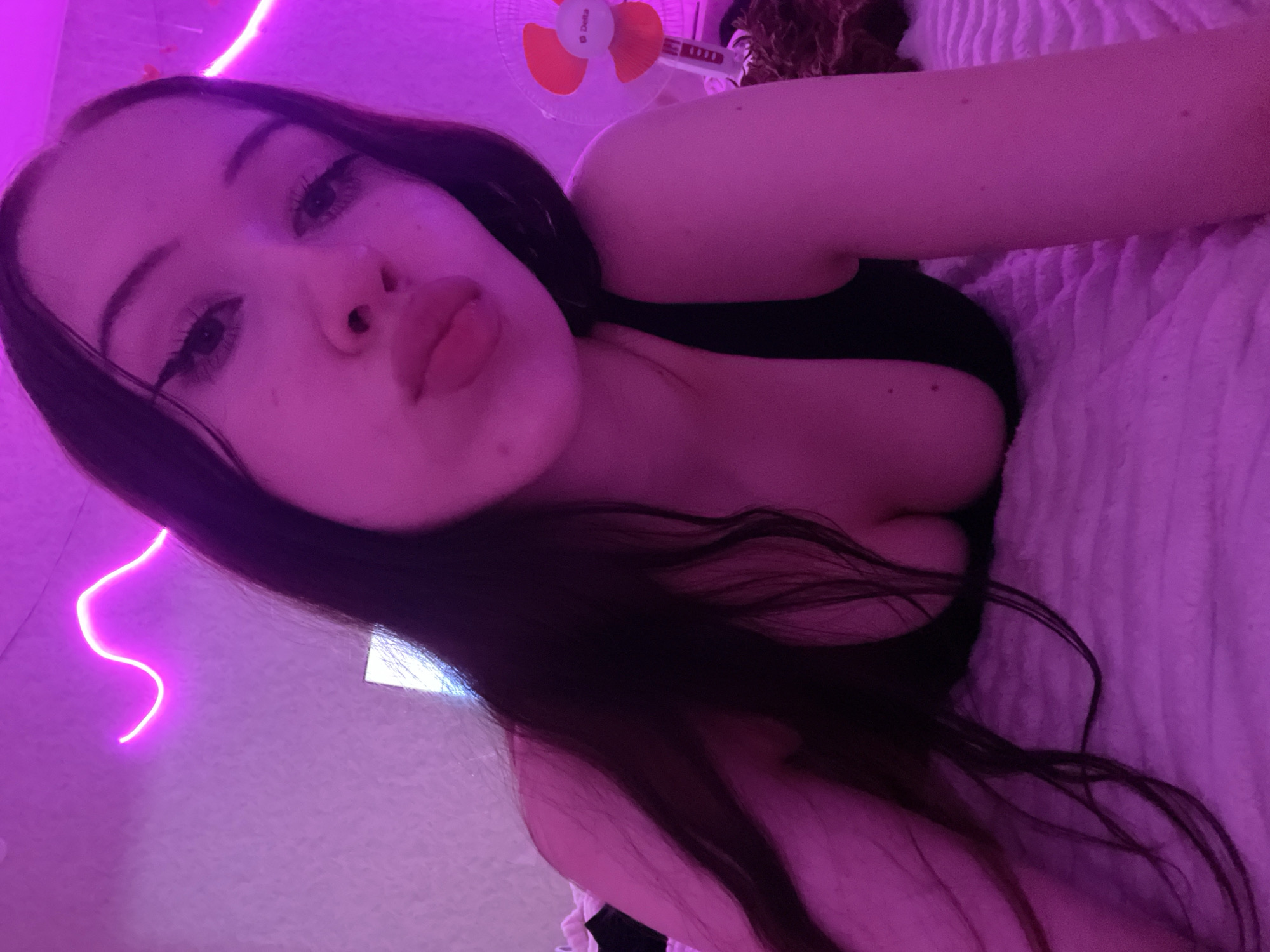 The image shows a young woman with long dark hair lying on a bed with a pink textured comforter. She is wearing a black top and has her lips puckered. The room is illuminated by pink neon lights, with a wavy design on the wall. A small white fan with orange blades is visible in the background to the right. The woman's face is close to the camera, with her eyes looking directly at it. The overall atmosphere is intimate and personal. There is no specific information within the image to indicate it is located in Votkinsk, Russia.