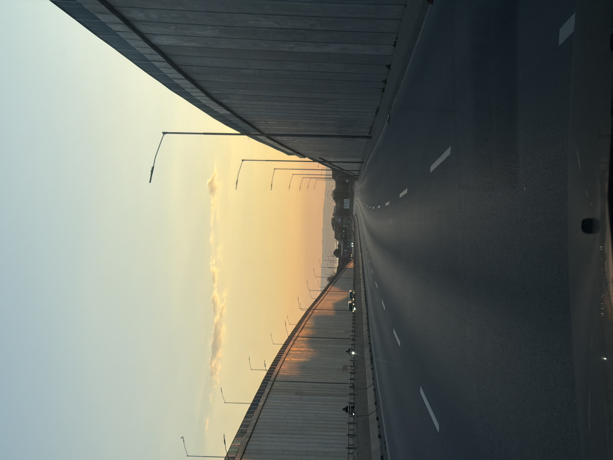 A long stretch of highway in Varna, Bulgaria, runs between two tall walls, likely part of a bridge or overpass structure. The sky in the distance is a gradient of light blue to orange, indicating either sunrise or sunset. Streetlights line the walls on both sides of the road, and occasional cars can be seen in the distance. The road surface is gray with white lane markings. The perspective suggests the photo was taken from inside a vehicle traveling along the highway.