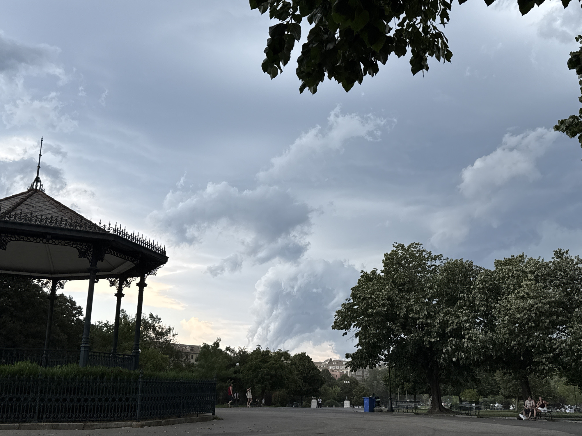 This media file depicts a park scene in Corfu, Greece, under a dramatic, cloudy sky. In the foreground, an ornate, dark metal gazebo stands to the left, fenced off by a black metal railing and manicured hedges. The gazebo has a red-tiled roof and a tall, slender spire.

To the right of the gazebo, a wide paved area suggests a promenade or park path. Several people are visible, scattered throughout the park. A couple sits on a bench beneath a large, leafy tree, while other individuals walk in small groups in the mid-ground. Beyond the path and trees, buildings with terracotta roofs are visible in the distance, hinting at an urban or semi-urban setting. Two large trees dominate the right side of the frame, their dense foliage contrasting with the lighter sky. A blue trash bin is also visible near the trees.

The sky is filled with large, imposing storm clouds, creating a sense of impending weather. The lighting is subdued, suggesting either early morning or late afternoon, possibly before or after a rain shower. The overall mood is somewhat somber due to the dramatic sky, but the presence of people enjoying the park suggests a moment of calm before potential weather changes. No visible text is present in the image.
