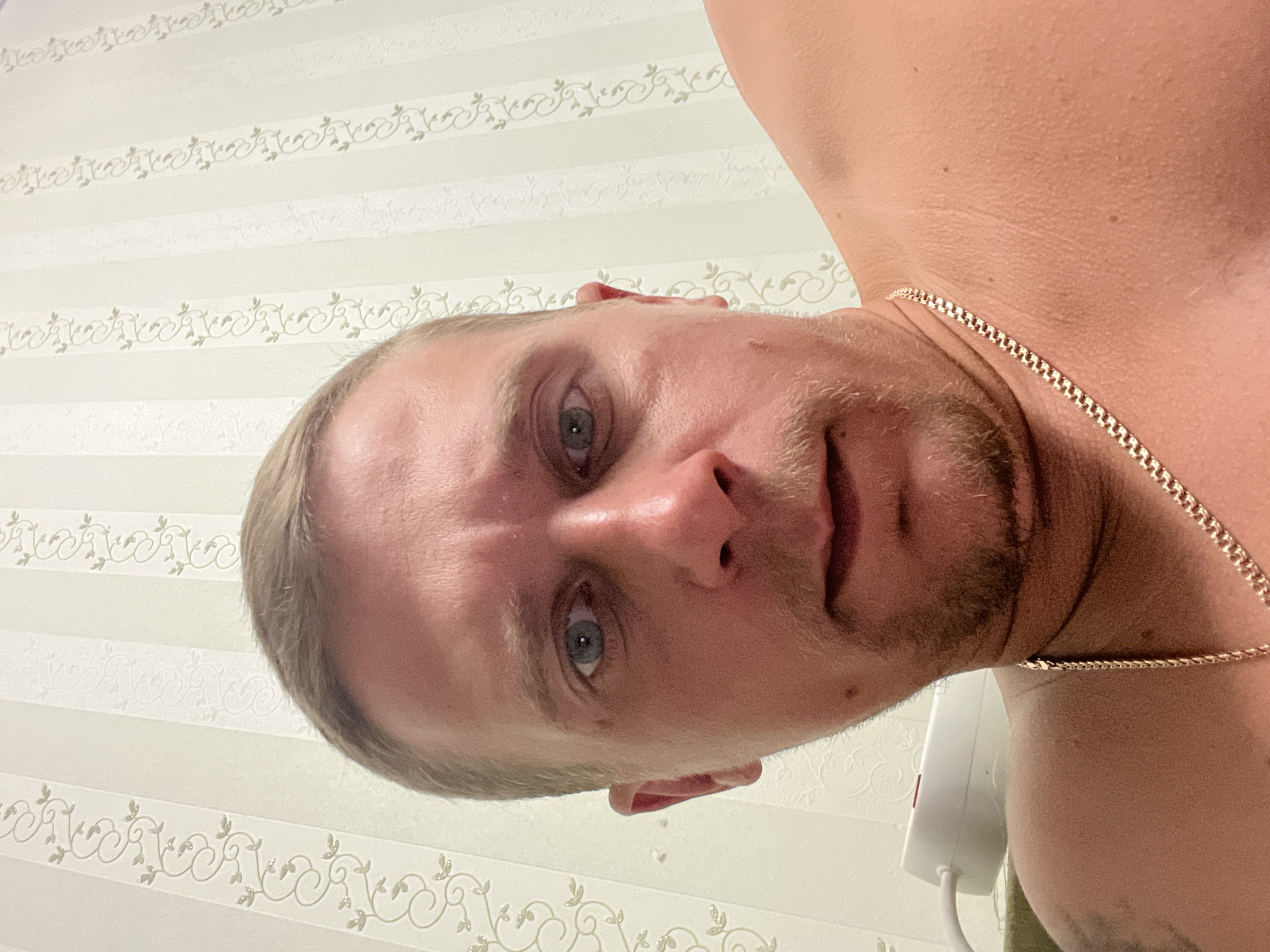 The image is a close-up, low-angle selfie of a Caucasian male with short blond hair and light blue eyes. He has a light beard and mustache, and is wearing a gold chain necklace. The background is wallpaper with vertical stripes and a floral pattern. To the left, a white electrical outlet strip with a cord is visible. The scene appears to be indoors, possibly in a bedroom, given the wallpaper. No specific location in Minsk, Belarus can be determined from the image alone.