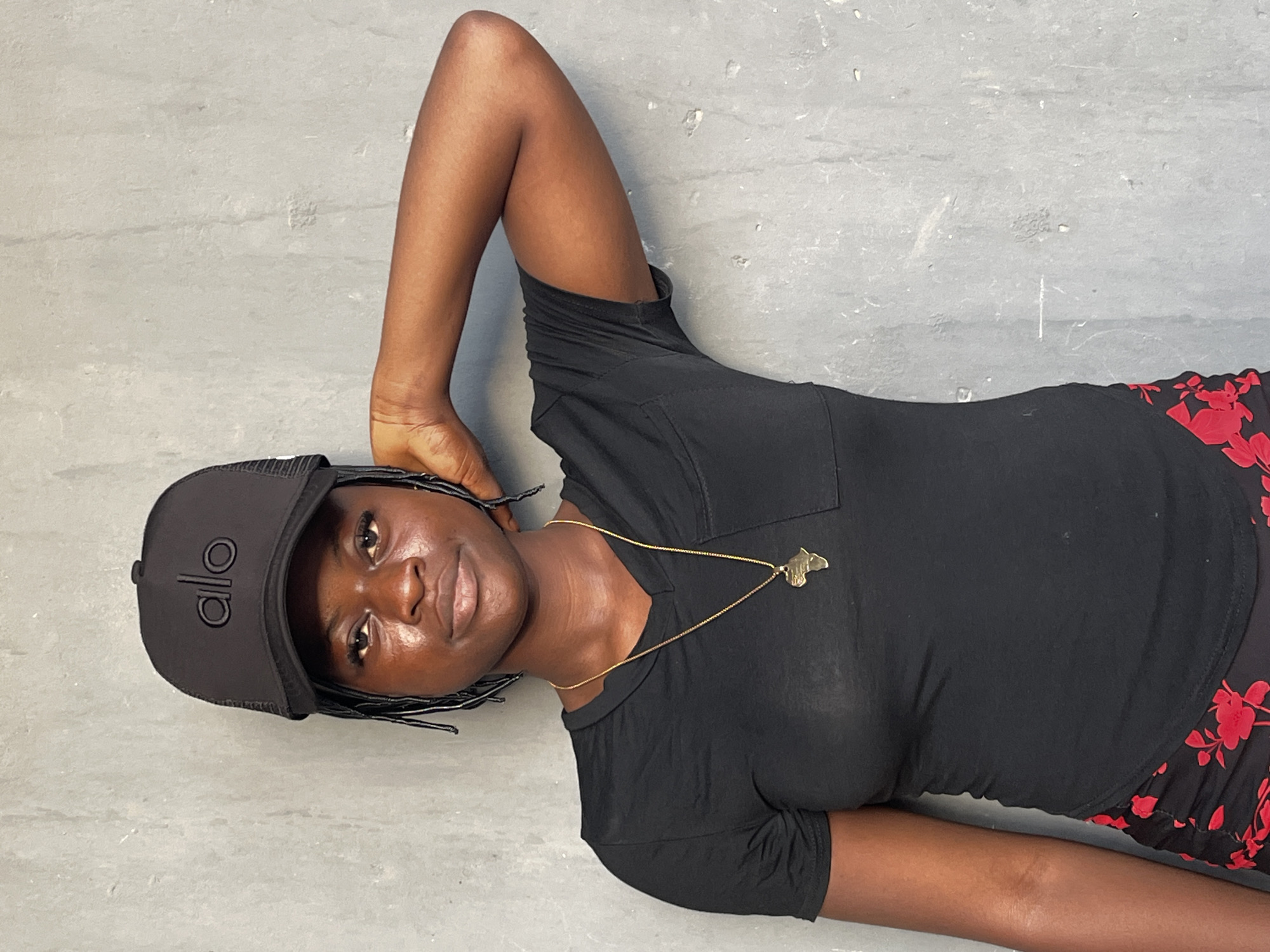 A woman lies on a gray surface. She wears a black "Alo" cap, a black t-shirt, and a red and black floral pattern bottom. A gold necklace with an Africa-shaped pendant is visible. Her right arm is raised, with her hand near the cap. The location is not identifiable as Badagry, Nigeria.