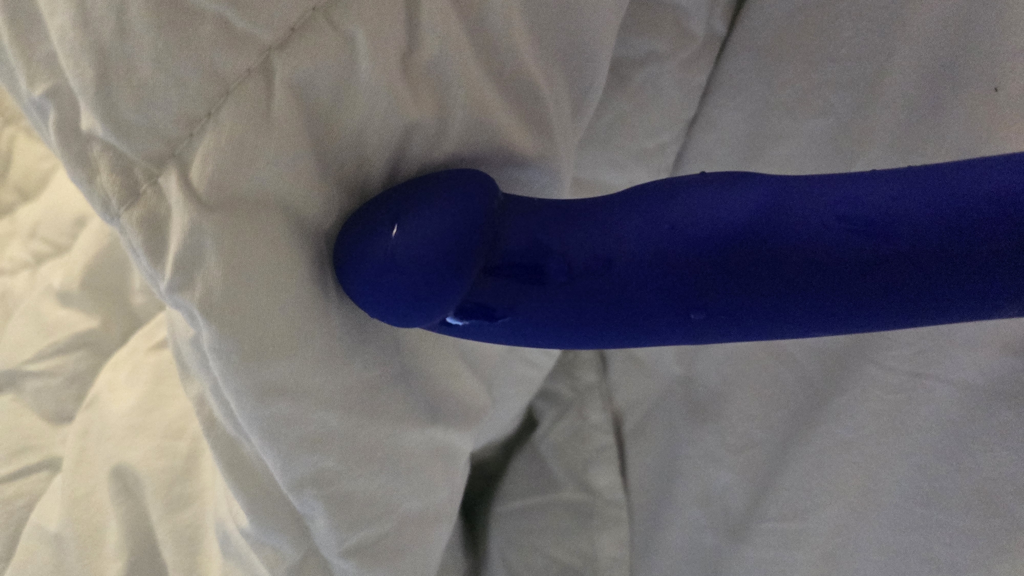 A close-up, low-angle shot shows the head of a blue vibrator peeking out from under a white duvet. The vibrator is wet, with visible droplets of liquid on its surface. The background consists of the white bedding, which is wrinkled and textured. The lighting is dim, creating shadows on the fabric. No location in Batumi, Georgia can be determined from the image.