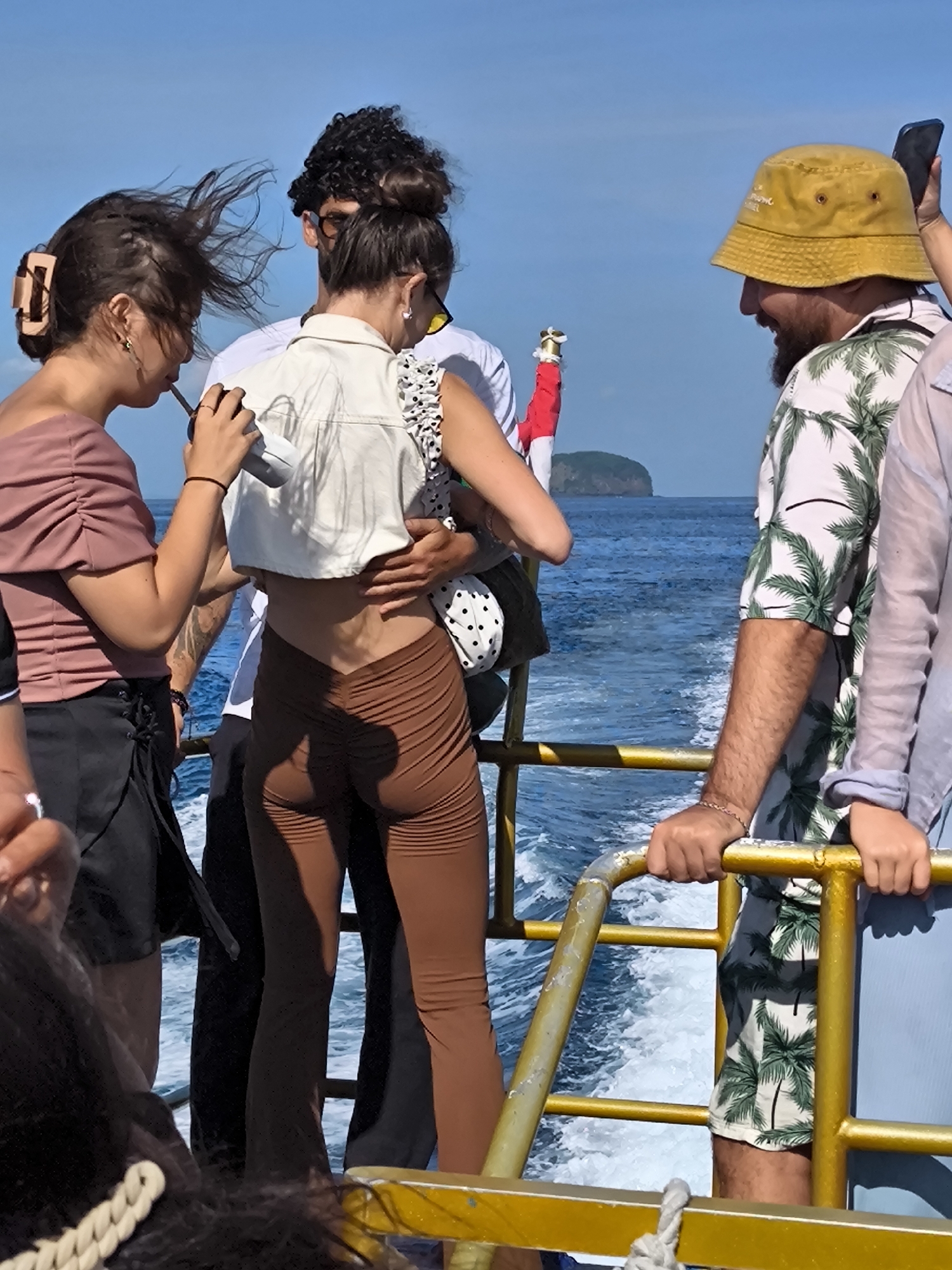 The image shows a group of people standing on a boat, possibly a tour boat, with the ocean visible in the background. The location appears to be coastal, with a small island in the distance. The sky is clear and blue.
From left to right:
- A woman with dark hair is wearing a pink off-the-shoulder top and black shorts. She is holding a cup with a straw and looking towards the center of the group.
- A man with curly hair and sunglasses is standing next to her.
- A woman in the center is wearing brown leggings, a white crop top, and a baby carrier with a child in it. She is being embraced by another person.
- A man with a beard and a yellow bucket hat is wearing a tropical-print shirt and is looking at the people to his left.
- Another person is partially visible on the right.
The boat has a gold-colored railing. The wake of the boat is visible in the water.