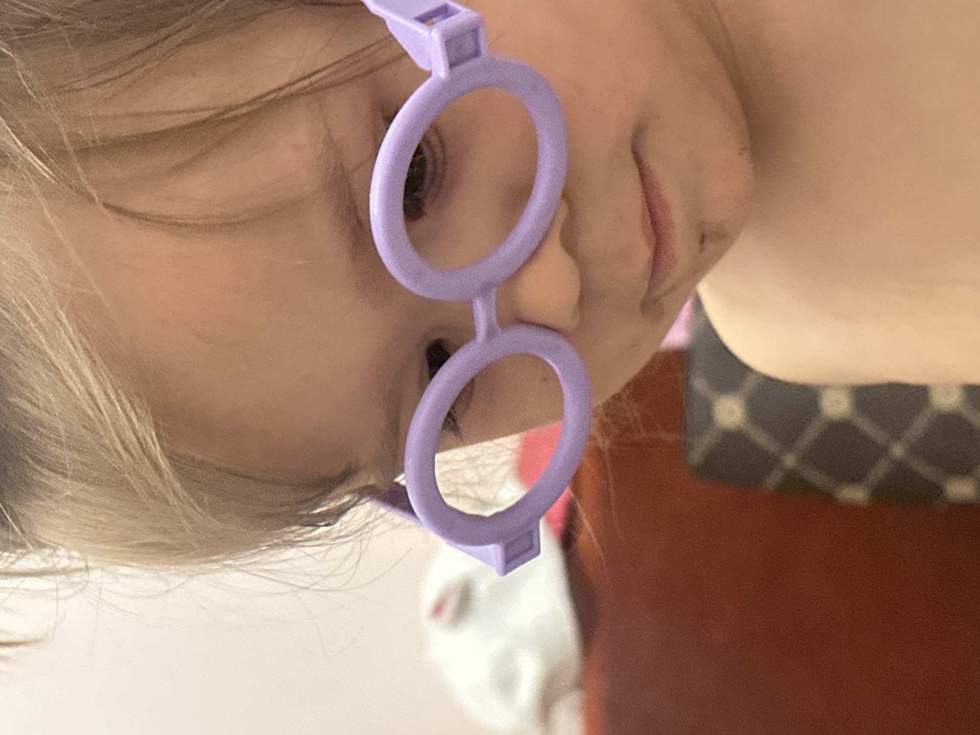 A close-up photograph depicts a child wearing toy glasses. The child has fair hair and light skin. The glasses are a light purple color and consist of two circular frames connected by a bridge. The background is blurry but appears to include parts of a patterned cushion and a red surface. The scene's location is unspecified.