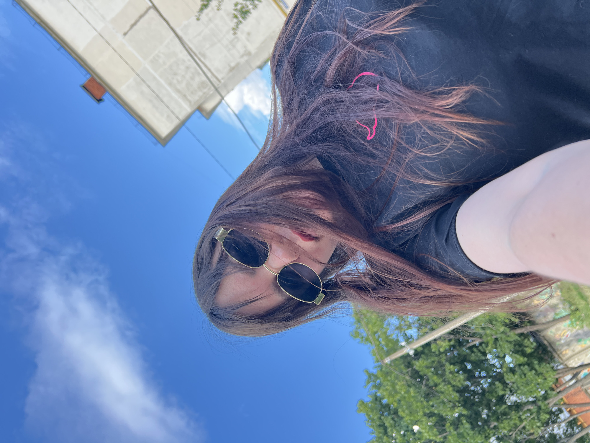 A young woman with long, dark brown hair, wearing sunglasses and a black t-shirt with a pink graphic, is positioned in the foreground of an outdoor urban scene. She is looking at the camera, and her face is partially obscured by her hair and sunglasses. The background includes a bright blue sky with wispy clouds, a portion of a light-colored multi-story building with a red brick chimney, and green trees. A utility pole is visible to the left of the woman. The image appears to be taken from a low angle, possibly a selfie. The location is identified as Noviy, Russia.