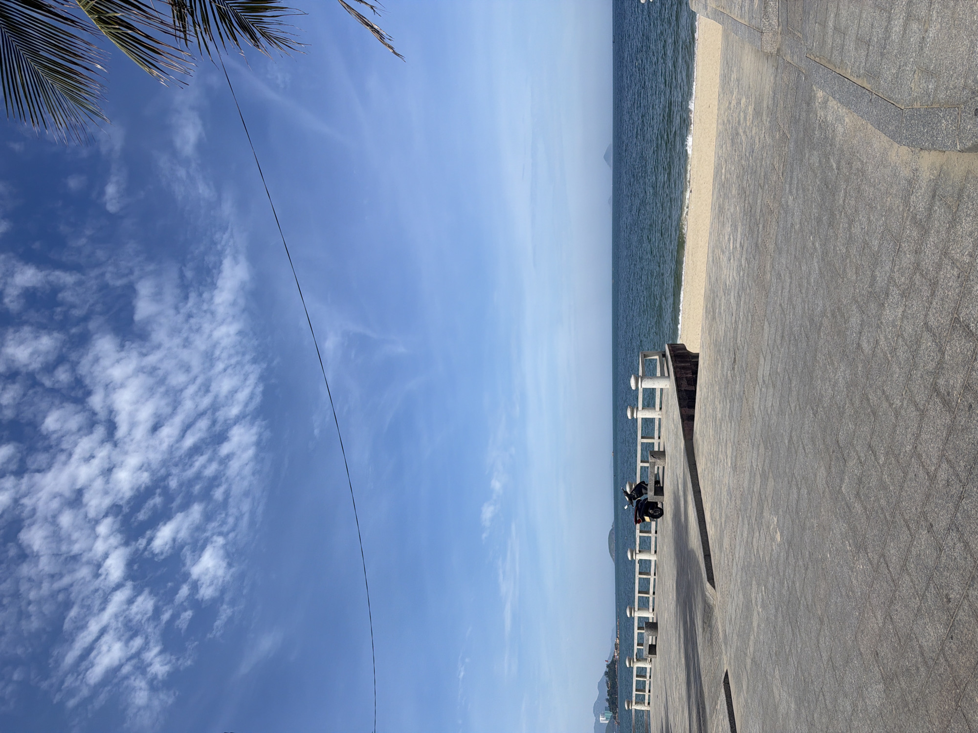 The image captures a serene coastal scene in Nha Trang Ward, Vietnam. A paved walkway, laid with grey stone tiles, occupies the foreground. To the left, a white railing runs along the edge of the walkway, overlooking a sandy beach. Beyond the beach, the calm blue ocean stretches towards the horizon, with a few small islands visible in the distance. The sky above is a clear blue, dotted with scattered white clouds. A single black scooter is parked on the walkway near the railing. Palm fronds are visible at the top right of the frame, suggesting the presence of palm trees nearby. The overall atmosphere is peaceful and tranquil, indicative of a quiet day by the sea.