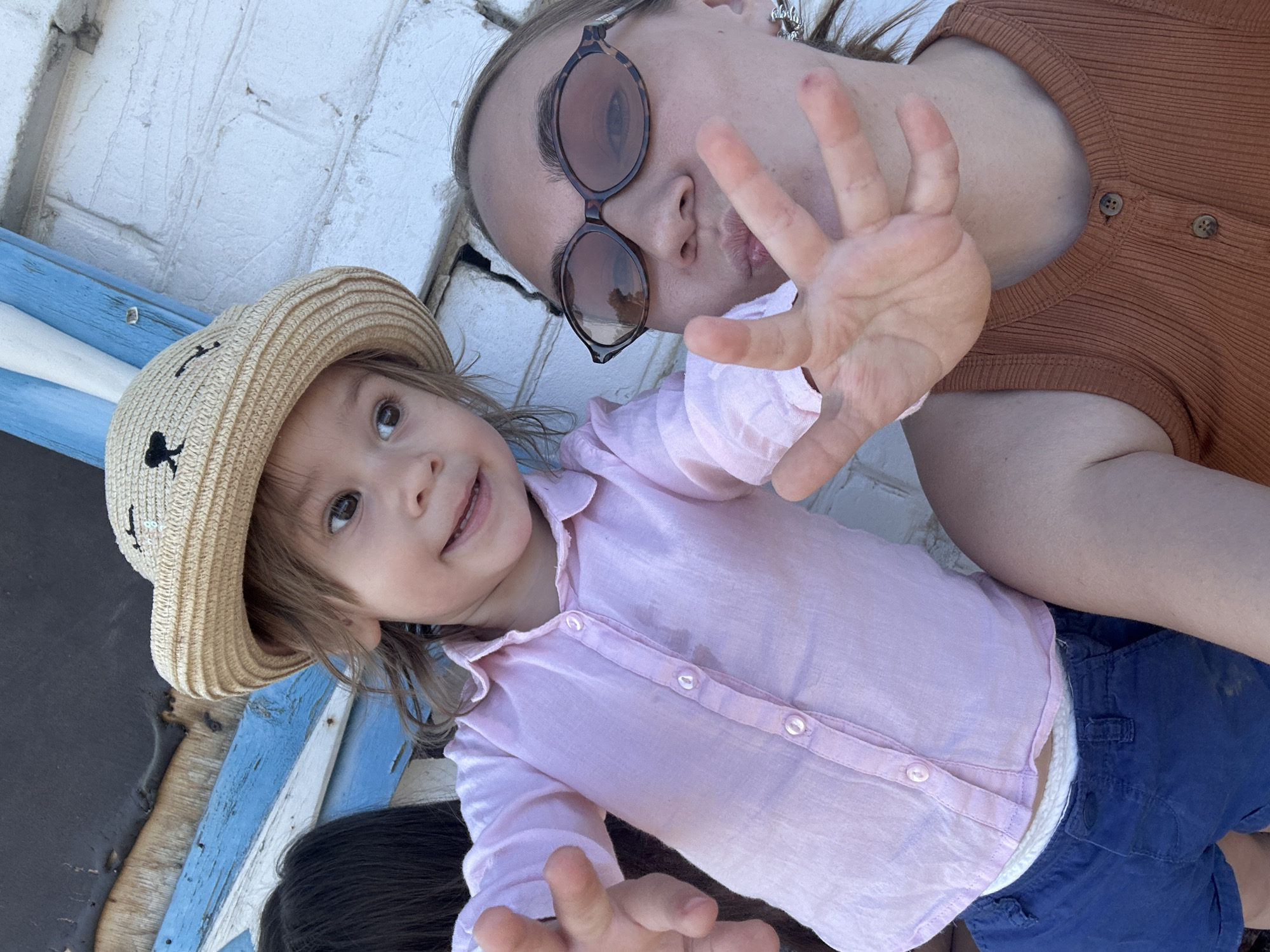 A woman wearing sunglasses and a brown ribbed shirt is lying down next to a young child with a straw hat and a light purple button-down shirt. The child is wearing blue shorts. The woman has her hand raised towards the camera. The background appears to be a white brick surface with blue and white painted wood panels. The image was taken in Chaikovsky, Russia.