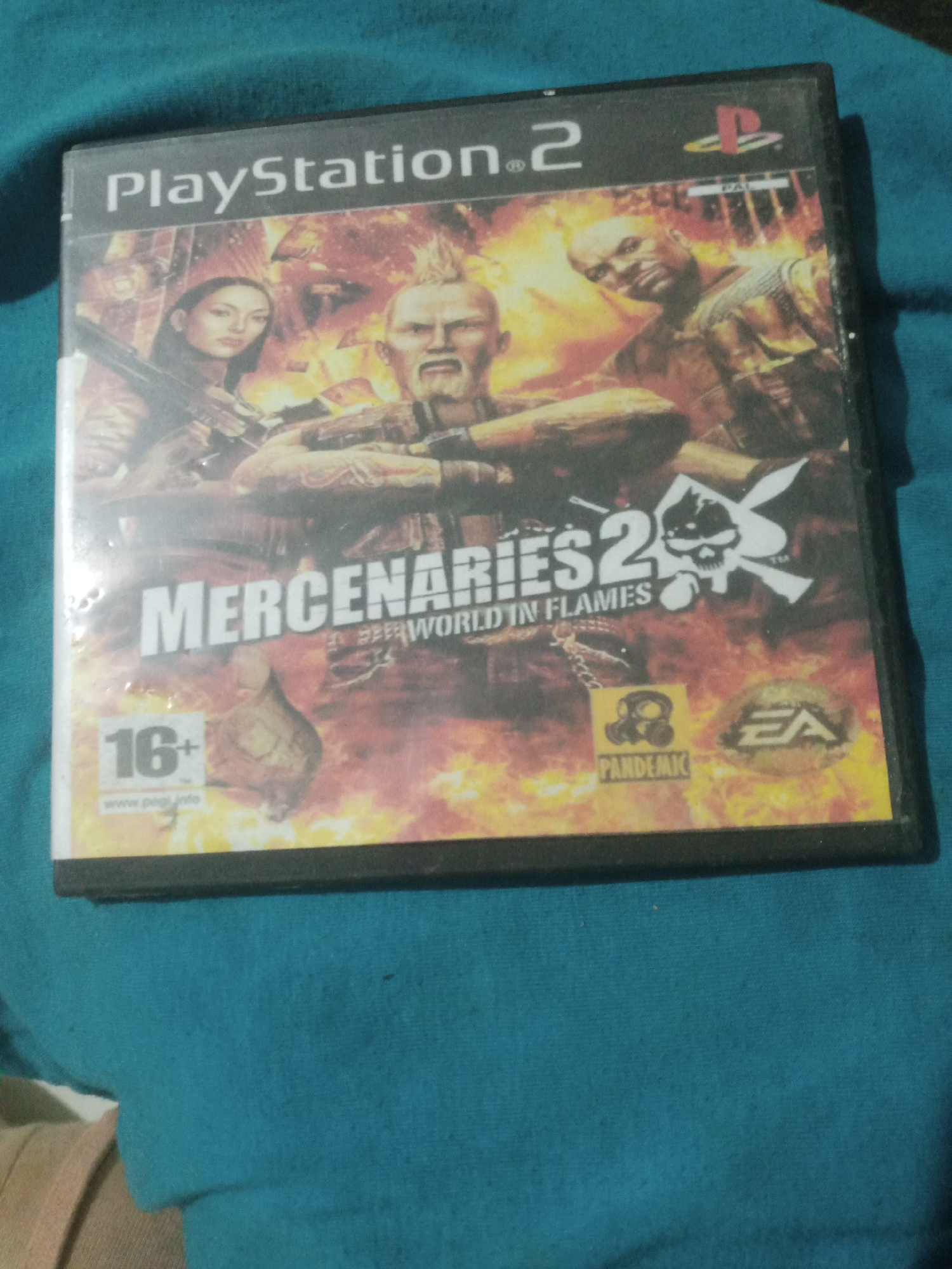 This is a close-up, eye-level, medium shot of a PlayStation 2 game case for "Mercenaries 2: World in Flames". The game case is resting on a textured teal fabric surface. The cover art depicts three characters in a fiery, chaotic environment, with flames and smoke prevalent. The "PlayStation 2" logo is visible at the top left, with the PlayStation symbol to its right. The game title "MERCENARIES 2" is prominently displayed in large white text. Below this, the subtitle "WORLD IN FLAMES" is visible. To the right of the title, a stylized skull logo with wings is present. In the lower left corner, a "16+" age rating symbol is shown, along with the website "www.pegi.info". To the lower right, there is a yellow square with a gas mask icon and the text "PANDEMIC", and an EA logo. The game case has some scuff marks and smudges, indicating it is used. The background is uniformly teal fabric with no discernible city elements. There is no information in the image that would allow for identification of a specific location within Santa Ana, Venezuela.