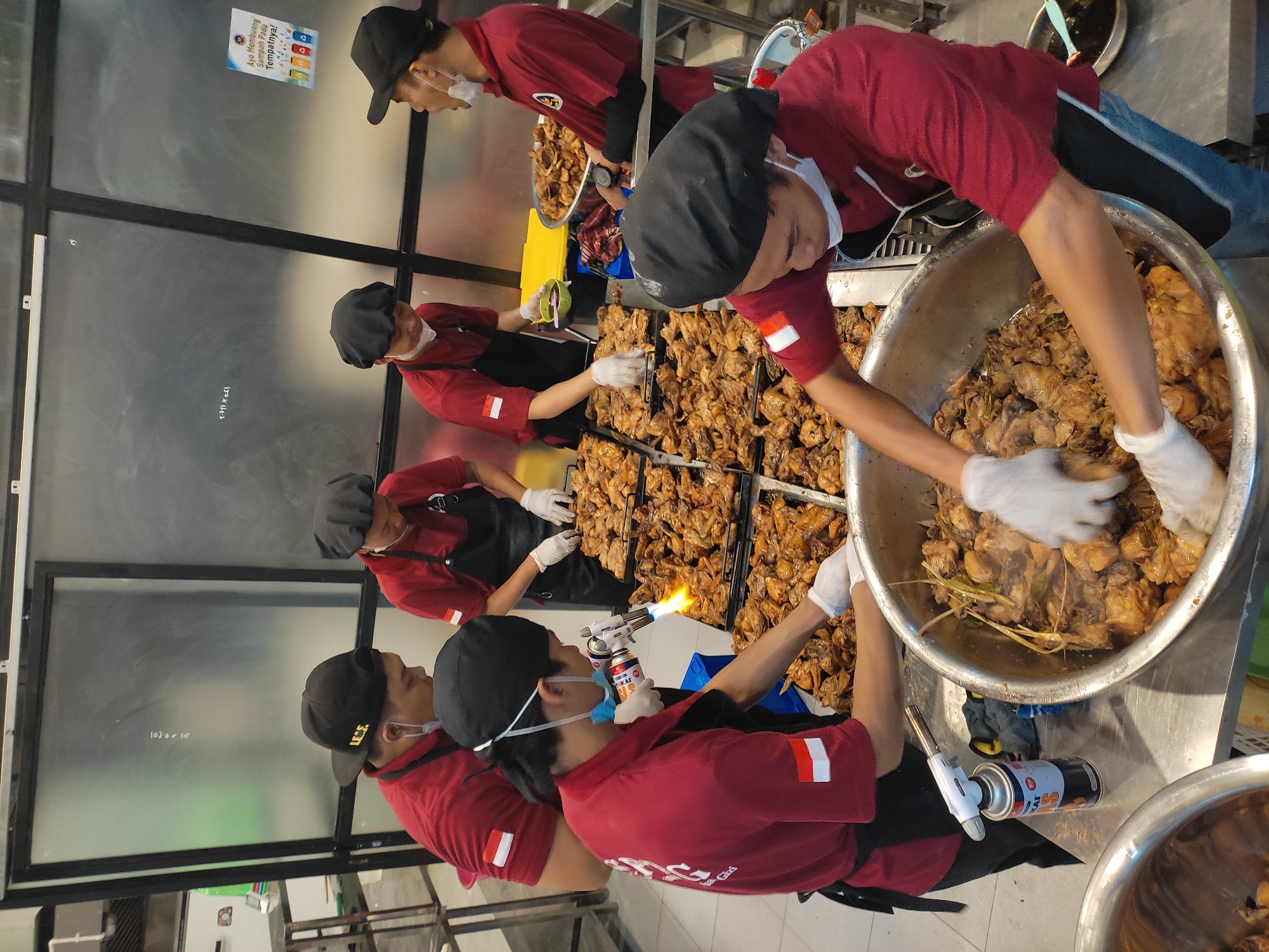 Six individuals, appearing to be kitchen staff, are engaged in food preparation within a commercial kitchen environment. They are all wearing red shirts with black aprons and black hair coverings. The individuals are in various stages of preparing large quantities of cooked chicken. One individual is using a culinary torch to caramelize or brown the chicken. Another individual is stirring chicken in a large metal bowl with white gloved hands. Trays of cooked chicken are spread across metal tables. The overall setting suggests a catering or food production facility. The presence of Indonesian flags on the sleeves of some of the individuals' shirts indicates their nationality. The backdrop includes metal shelving units and what appear to be industrial kitchen appliances, including a large exhaust hood system. A sign in Indonesian, related to waste disposal, is visible on a glass partition. While the specific location in Ponorogo, Indonesia is not explicitly depicted, the context of food preparation and the national flag suggest a connection to the region.
