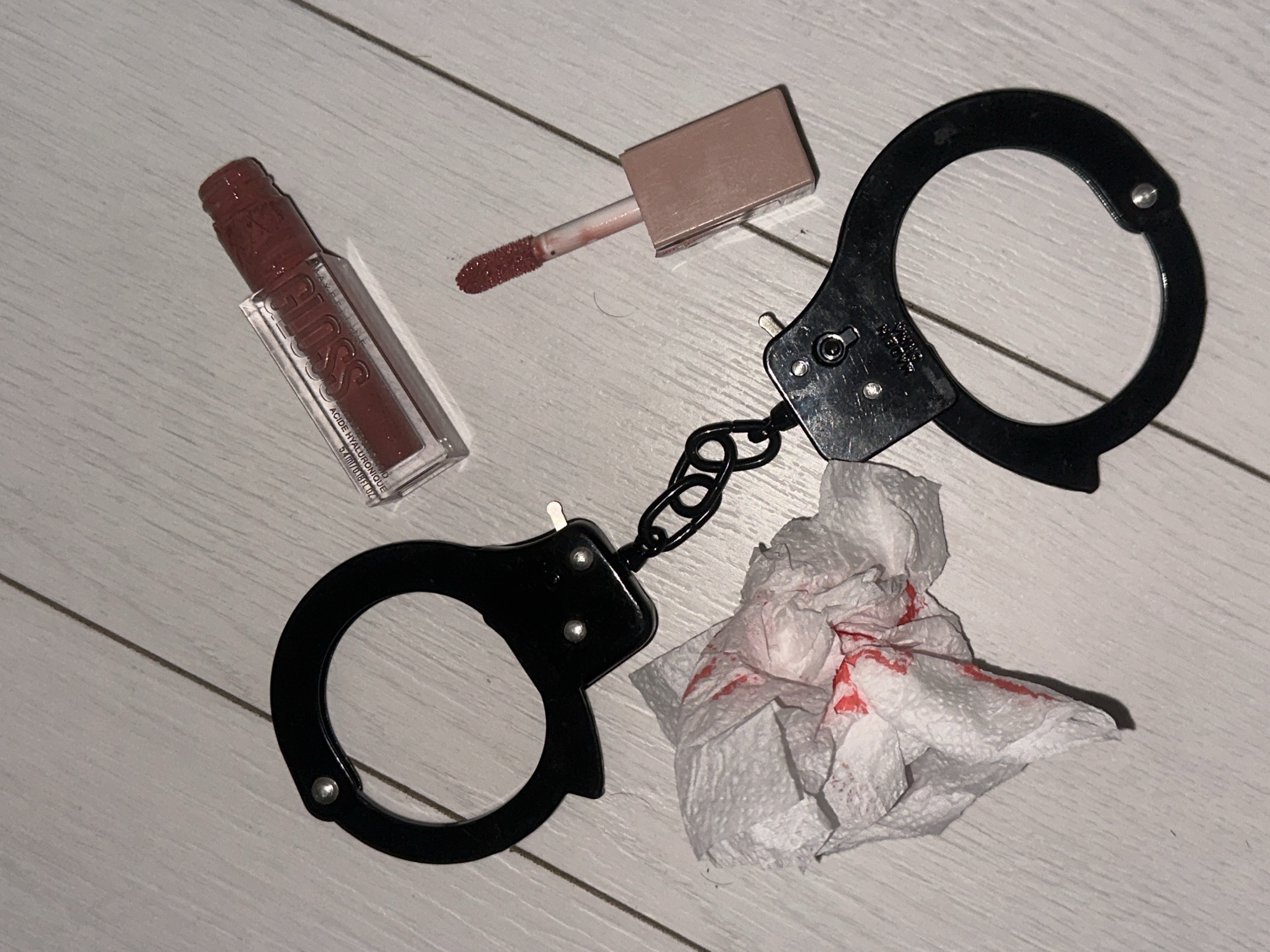 The image depicts a collection of items scattered on a light-colored wooden floor, possibly a bedroom or bathroom. In the foreground, a pair of black, metal handcuffs are partially open. Adjacent to the handcuffs is a crumpled white tissue with streaks of red substance, possibly lipstick or blood. To the upper right, a clear tube of lip gloss with a red cap lies on its side, with a small amount of the product also smudged on the floor. Beside it, the applicator wand from the lip gloss is positioned upright, its tip coated in the same red substance. The overall arrangement suggests a personal, perhaps dramatic or humorous, scene. There are no indications of a specific location in Sofia, Bulgaria, within the image itself.