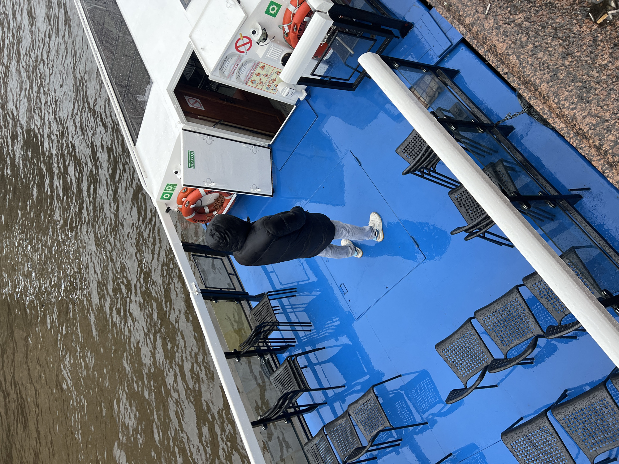 A high-angle shot depicts the open deck of a tour boat on a body of water in Saint Petersburg, Russia. The deck is painted a vibrant blue and has rows of empty metal chairs. A person wearing a dark hooded jacket and light-colored pants stands in the center of the frame. Near the front, there is a white structure containing safety equipment like life preservers and emergency instructions. A white railing runs along the edge of the boat. Visible outside the boat is the murky water and a portion of a granite embankment. The seating and deck surface suggest it may be wet from rain.