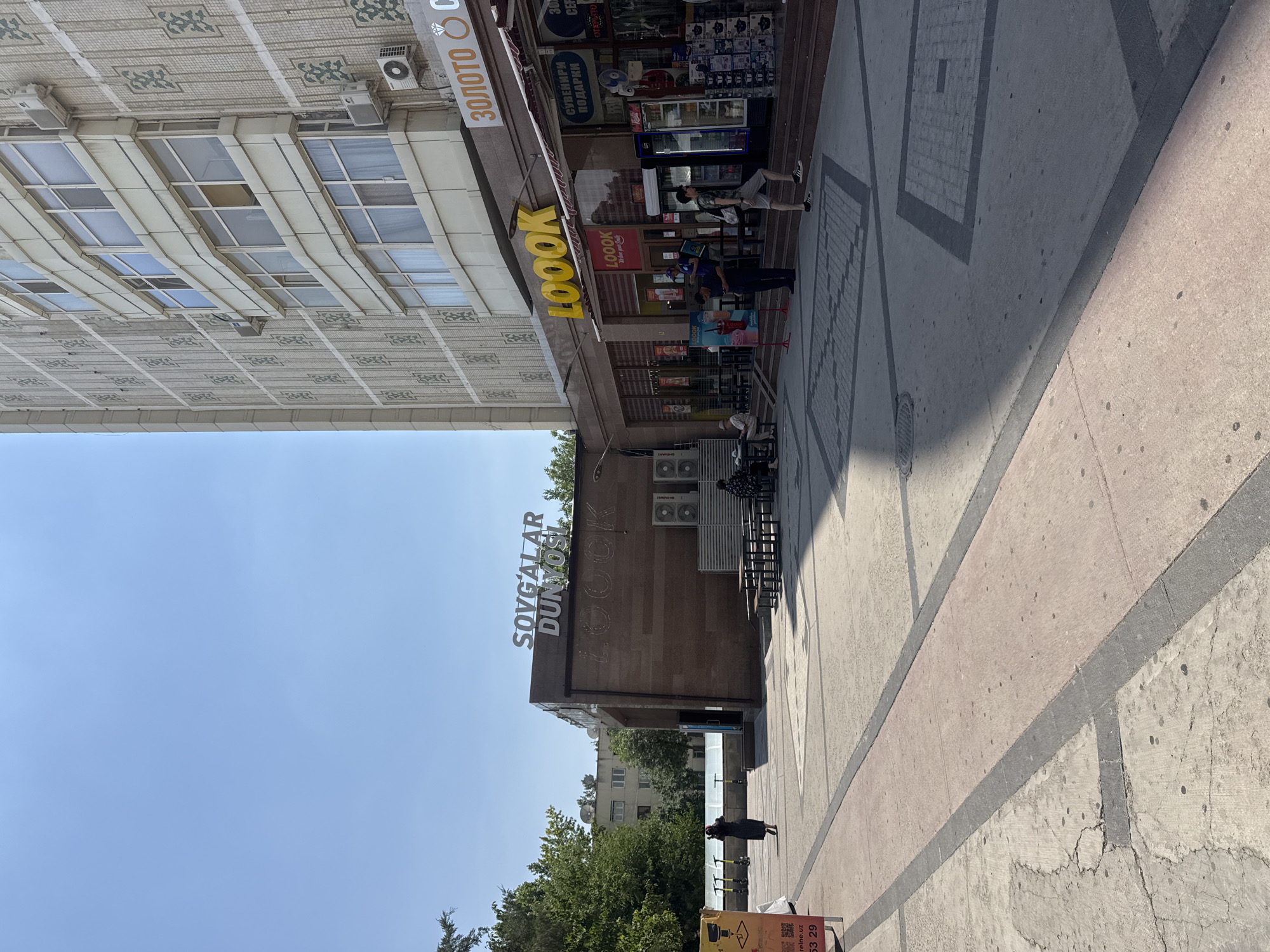 In an outdoor urban setting under a clear blue sky, a modern multi-story building with a patterned facade stands on the right, its cream-colored exterior accented with decorative green elements and numerous windows. Below the building, a commercial storefront displays a sign reading "LOOOK" in large yellow letters, beneath which is a smaller sign saying "LOOOK We love your Smile." Adjacent to this is a section with "ЗОЛОТО" and a ring symbol, suggesting a jewelry store, and another sign with "СУВЕНИРИ ПОДАРКИ" (Souvenirs Gifts). Several people are visible: a person in a dark dress walks away from the camera on the left; two people sit at an outdoor table on the lower right; a man in a camouflage shirt and shorts walks up stairs towards the stores; and two men in dark uniforms stand near the entrance of the LOOOK store. In the background, a lower, darker brown building has a sign with "SOVG'ALAR DUNYOSI" in white letters. The foreground features a paved area with distinct sections of gray and reddish-pink concrete, marked with darker gray lines. A few electric scooters are parked on the left side of the image.