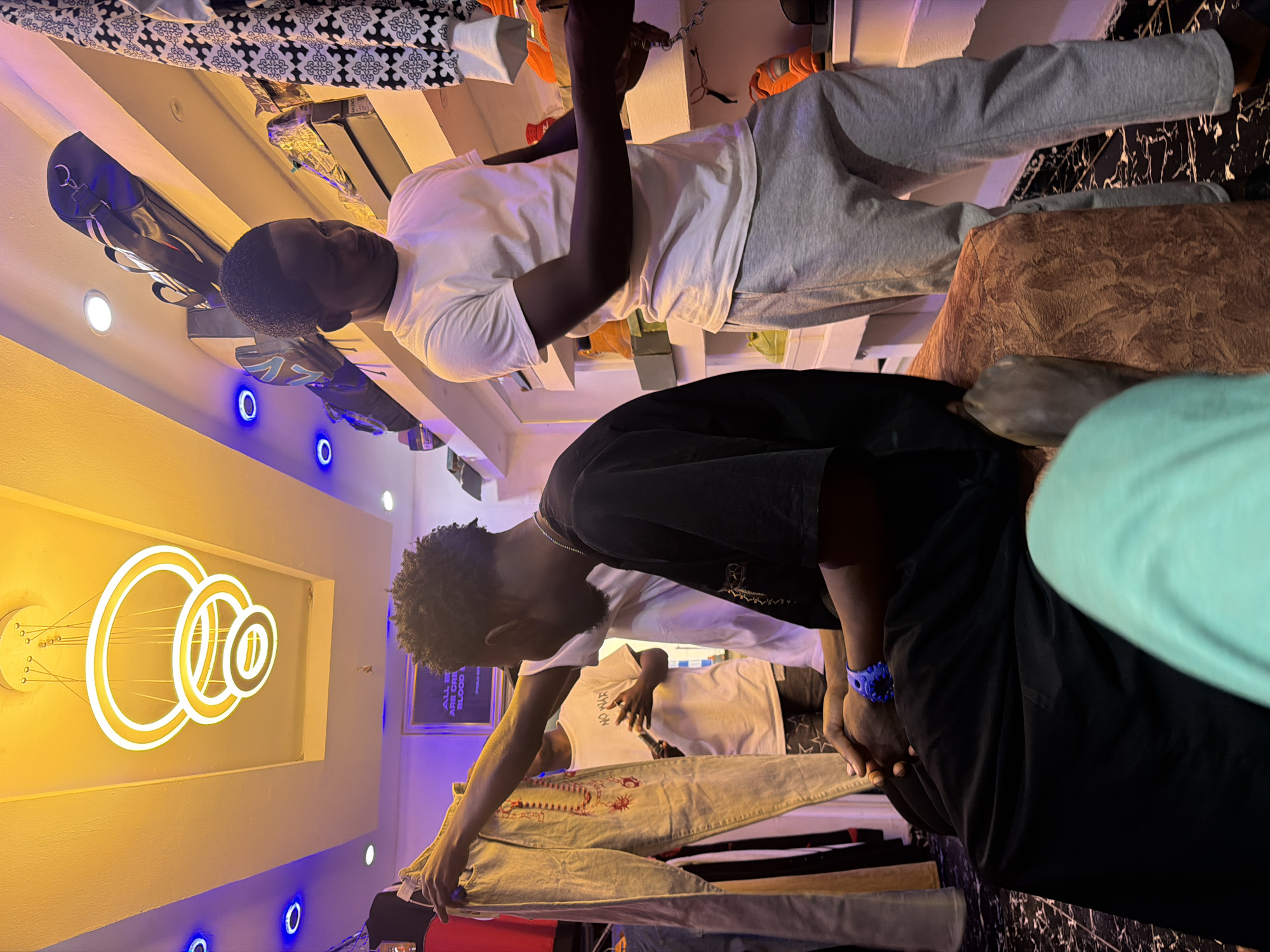 In a retail clothing store in Bukuru, Nigeria, a young Black man wearing a white t-shirt and gray sweatpants stands examining clothing. To his left, another Black man, wearing a black t-shirt, sits on a brown, textured ottoman, holding a pair of light-colored pants. A third Black man, wearing a white t-shirt with "NO WAR" printed on it, is visible in the background. The store's interior features a modern chandelier with multiple circular lights, and various clothing items are displayed on racks and shelves. The flooring is black and white tiled.