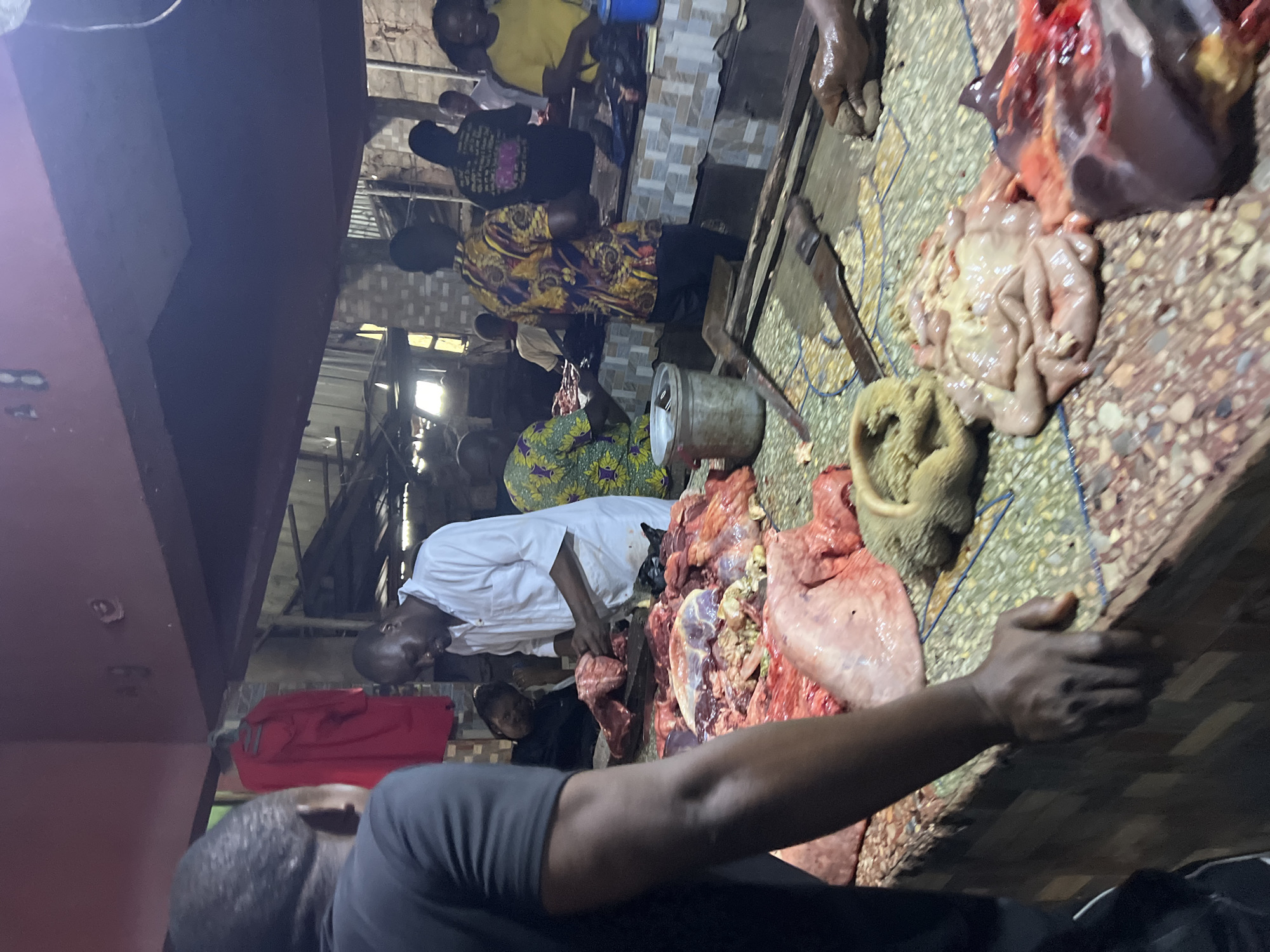 The image depicts a busy scene inside a market, likely in Nkpor, Nigeria. In the foreground, a large, butcher's counter is laden with raw meat and offal, including what appears to be organs and intestines. Several individuals are engaged in the sale and preparation of these items.
A man in a white shirt stands behind the counter, actively working with a piece of meat, suggesting he is a butcher. To his left, another person, partially visible, seems to be assisting or observing. In the background, more individuals are present, some tending to stalls and others engaged in transactions. The market environment is characterized by its rustic construction, with visible wooden beams and corrugated metal sheeting forming the roof. Red clothing is visible hanging in the background, adding a splash of color to the otherwise muted tones of the scene. The lighting appears to be a mix of natural light filtering in from openings and artificial sources, creating shadows and highlighting the details of the butchered meat. The overall impression is one of a lively and functional marketplace.