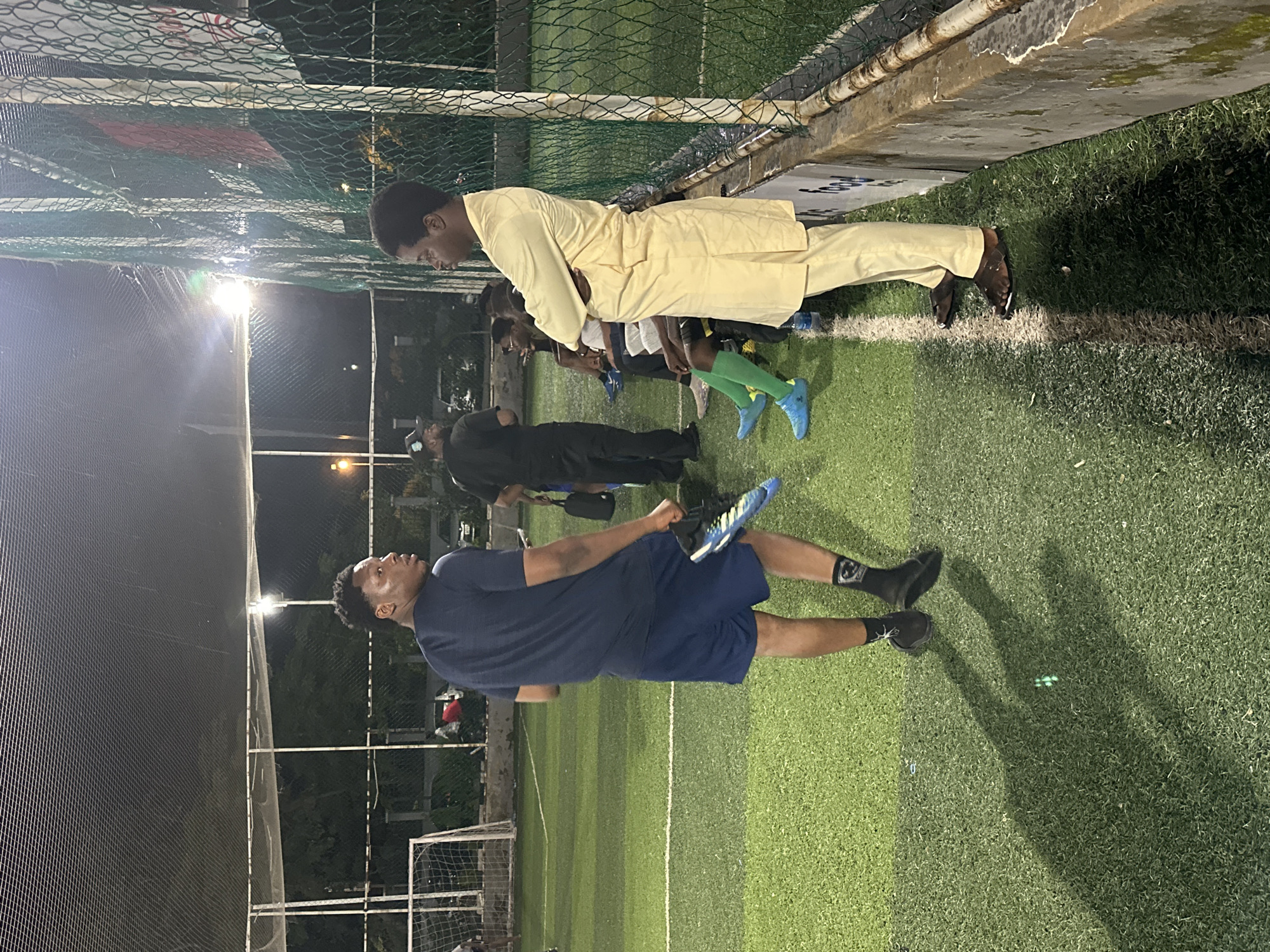 An outdoor football pitch in Abuja, Nigeria, is illuminated by floodlights at night. The artificial turf is marked with white lines. In the foreground, a young Black man wearing a dark blue t-shirt and blue shorts stands with his back to the camera, holding a pair of blue and black football boots. To his right, another young Black man in a light yellow kaftan and pants stands with his arms crossed, looking to the side. Behind them, several other young Black men are seated on a low wall or bench, some wearing football attire. The scene is enclosed by a high net fence. The lighting suggests a sporting event or practice session is taking place.