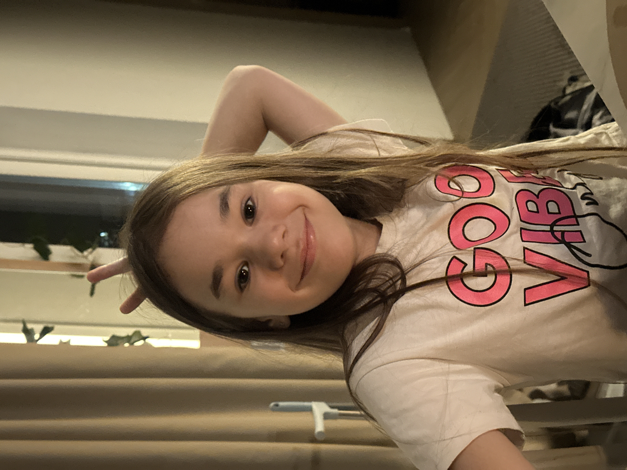 A young girl with long brown hair is pictured indoors. She is wearing a white t-shirt with pink lettering that says "GOOD VIBES". She is smiling and making a peace sign with her left hand near her head. The background appears to be a room with blinds partially visible behind her. The location is not detectable as the city of Kharnavy, Russia from the image.