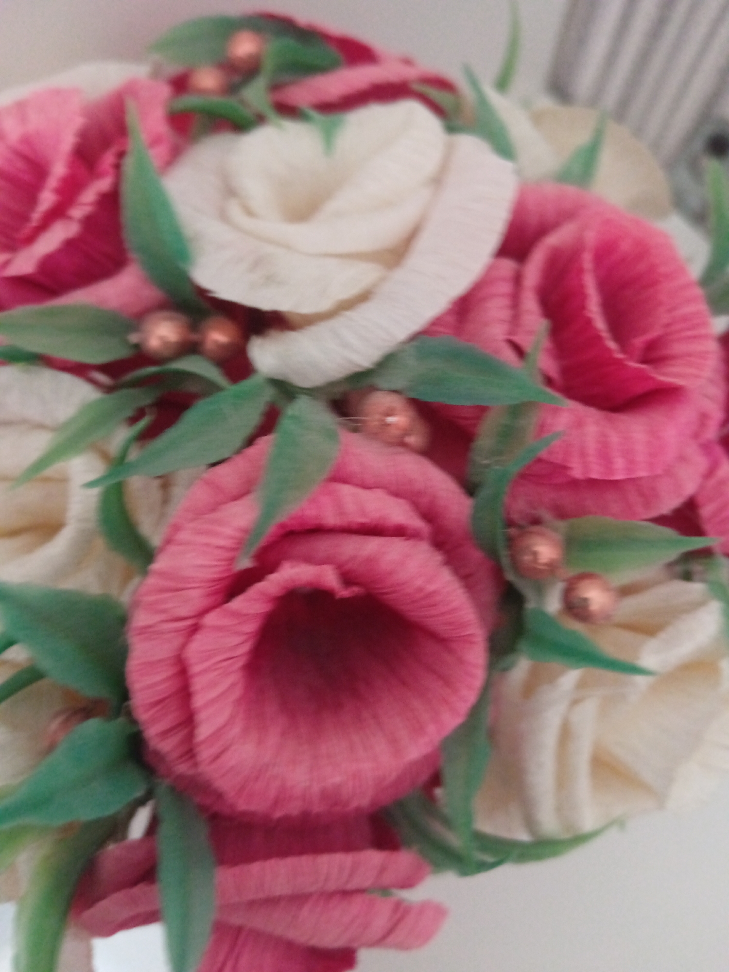 A close-up, overhead shot showcases a bouquet of artificial roses, crafted from crepe paper. The roses are predominantly pink and cream, interspersed with green paper leaves and adorned with small, copper-colored beads. The bouquet is arranged in a tight cluster, with some roses facing upwards and others angled to the side. The background is a soft, blurred off-white, suggesting an indoor setting. The image is slightly out of focus, with a shallow depth of field that blurs the edges of the bouquet and the background. No specific location in Sorochinsk, Russia, is identifiable from the image.