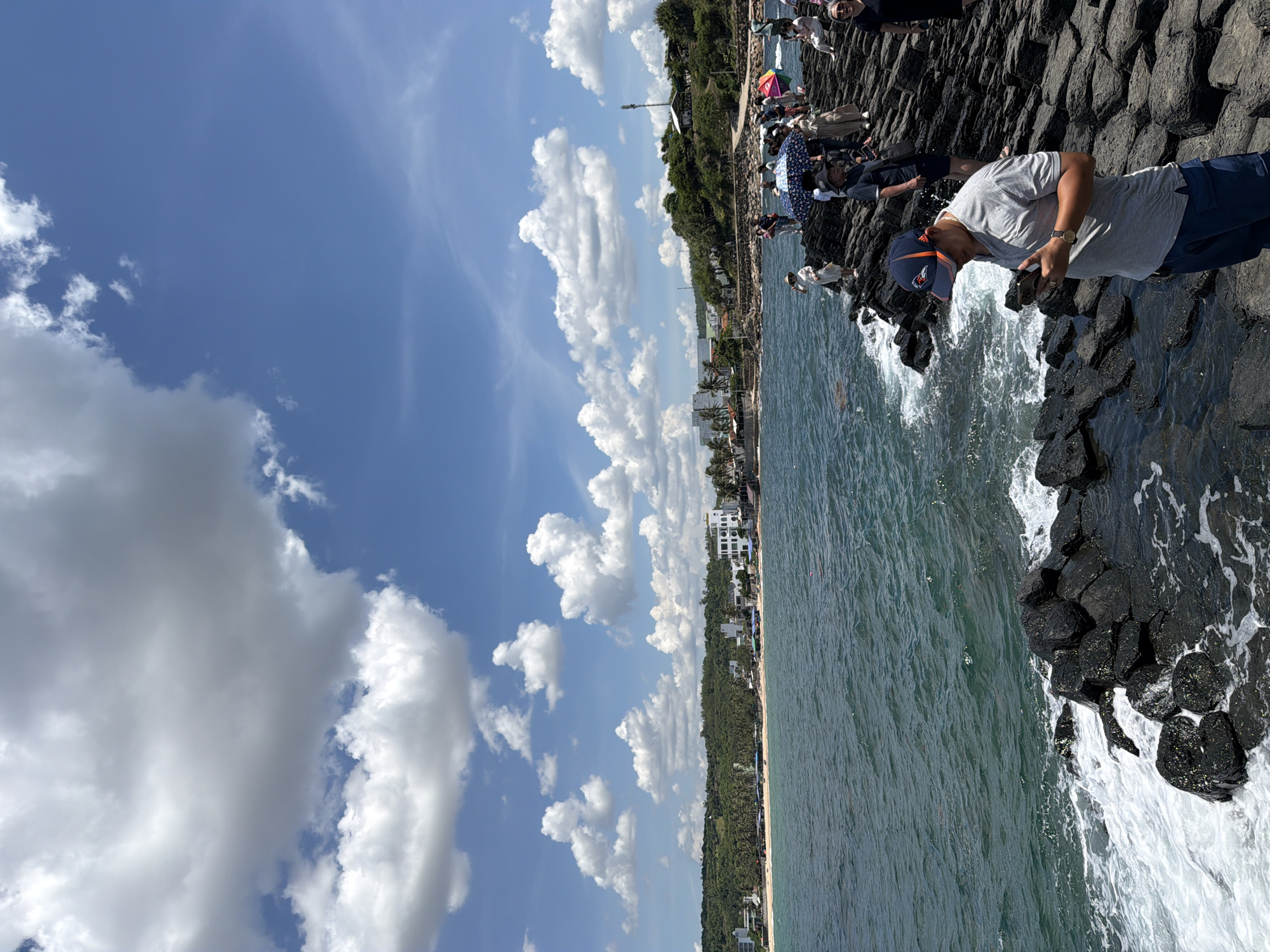 The image depicts a scene at Gành Đá Đĩa, located in the city of Phú Lương, Vietnam. The foreground is dominated by a man wearing a blue and orange baseball cap and a grey polo shirt, looking down at his mobile phone. He is standing on a formation of dark, columnar basalt rocks that extend into the choppy, blue-green ocean. Numerous other people are scattered across the rocks, some holding umbrellas, suggesting a tourist or recreational area. In the background, a coastline stretches out, featuring a sandy beach lined with palm trees and various buildings, including hotels and restaurants. The sky above is a vibrant blue, dotted with fluffy white clouds. The waves are crashing against the basalt formations, creating white foam.