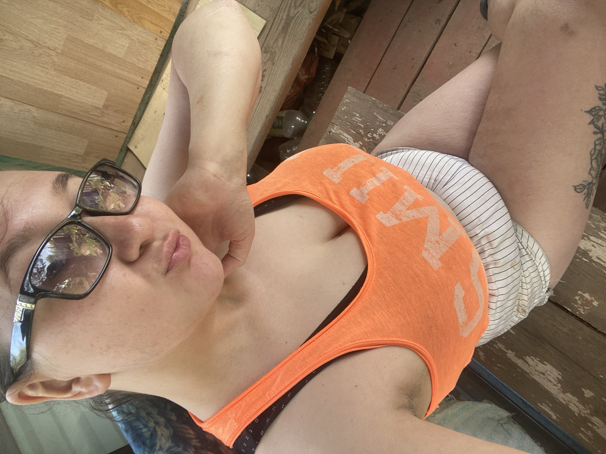 A woman lies on a weathered wooden surface. She wears glasses, an orange tank top, and striped shorts. A tattoo is visible on her upper thigh. The location is not discernable in the image, but is said to be in Прикамский, Russia. Bottles and other debris are visible in the background near the woman.