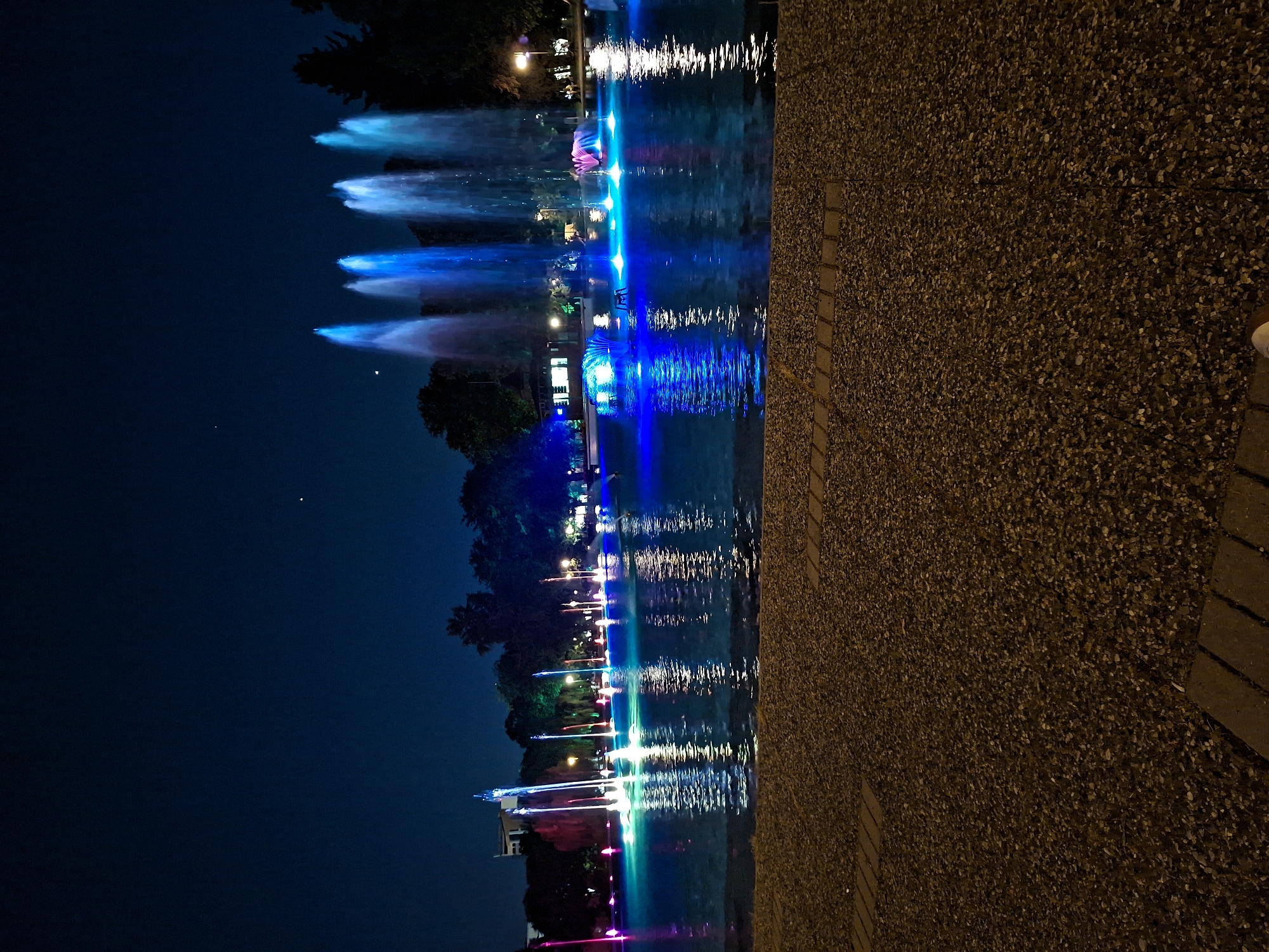 The image shows a night view of a water fountain display in Plovdiv, Bulgaria. The fountains are illuminated with colorful lights, primarily blue, green, white, and pink. The water jets are reflected in the surface of the water, creating a mirrored effect. Dark trees surround the area behind the fountain. To the right of the water feature is a walkway composed of small pebbles and brick border segments. The sky is dark, suggesting it's a night scene.