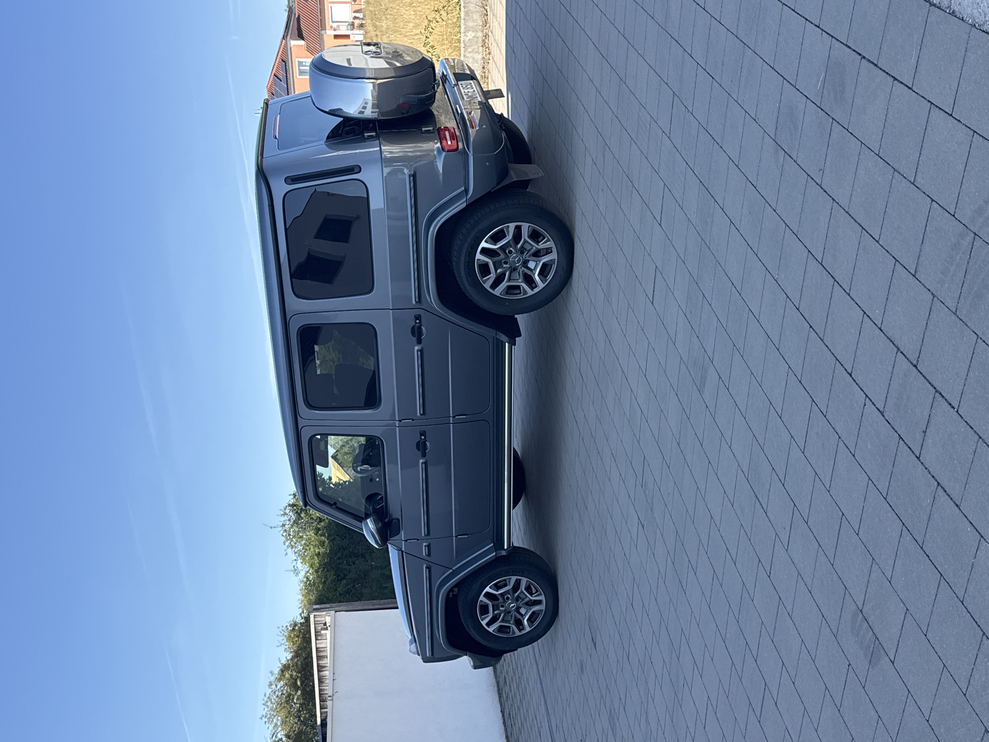In Wettstetten, Germany, a dark grey Mercedes-Benz G-Class is parked on a paved driveway under a clear blue sky. The vehicle is viewed from its rear quarter, showcasing its rugged design with a spare tire mounted on the back. The car's windows are tinted, and its license plate is partially visible. Behind the car, a low hedge and a portion of a building with an orange roof are visible. The driveway is composed of rectangular grey pavers arranged in a staggered pattern. The scene suggests a residential setting on a bright day.