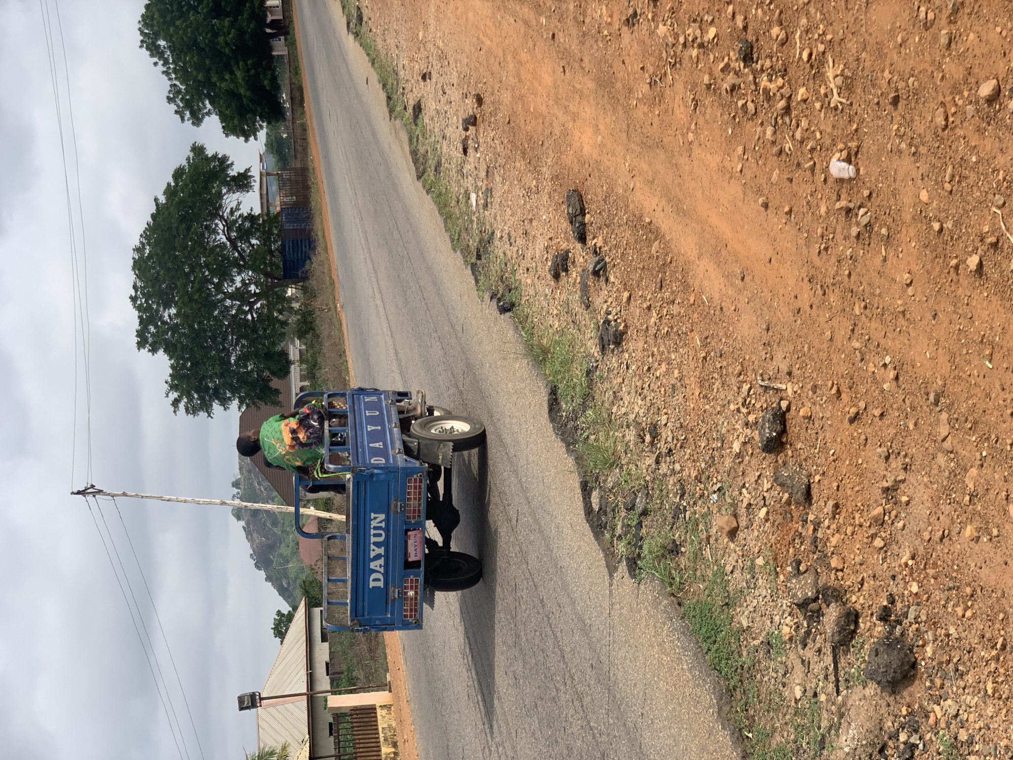 The image shows a blue "DAYUN" tricycle on an asphalt road, likely in Kabba, Nigeria. A person wearing a green shirt is seated in the tricycle. The road is bordered by dirt with sporadic vegetation on the right. Debris, including dark organic matter and a discarded plastic bottle, is scattered along the road's edge. In the background, buildings, trees, a utility pole, and a hill are visible. The sky is overcast.