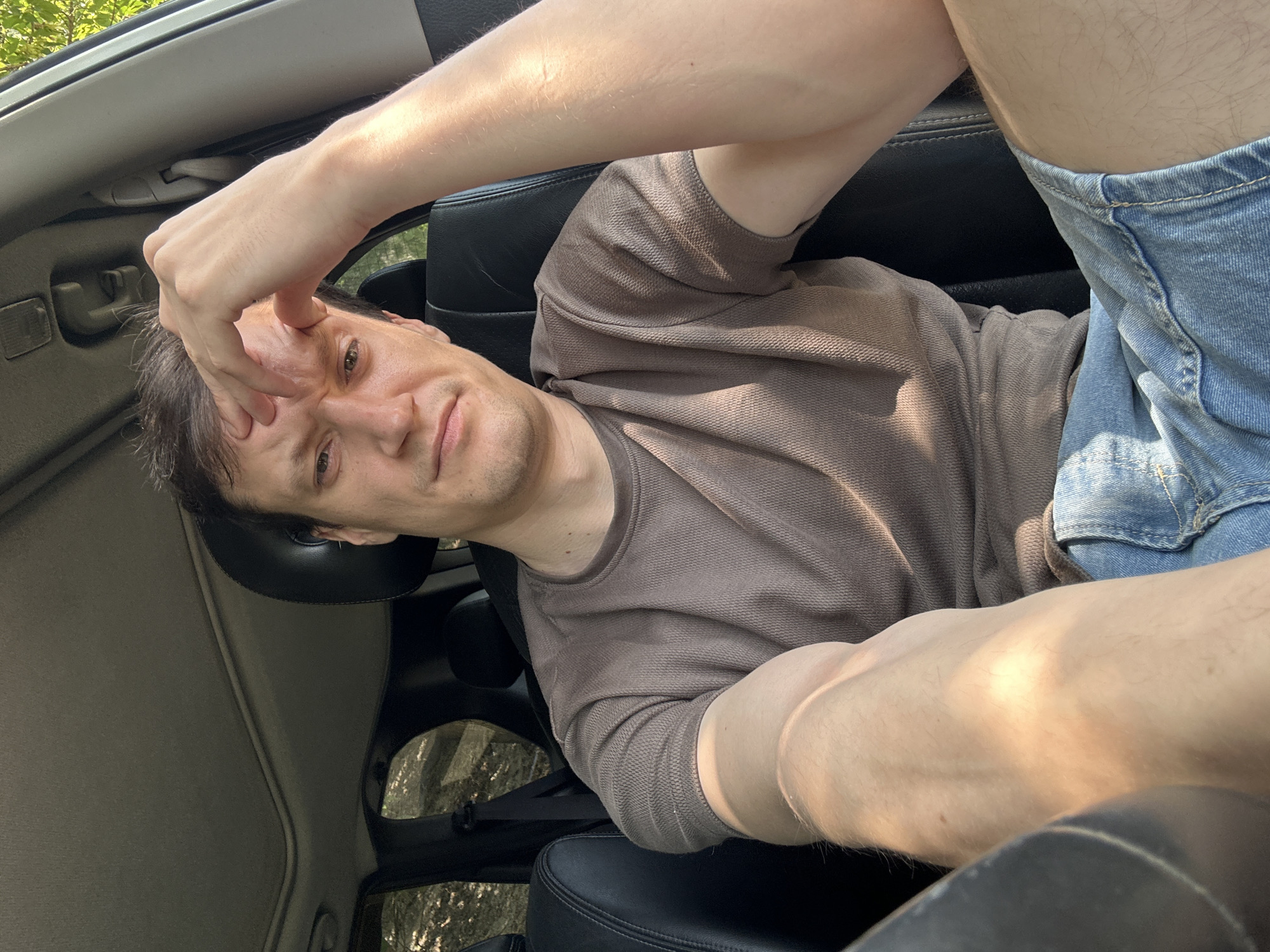 A man is sitting in the driver's seat of a car, possibly a SUV due to the sunroof visible in the image. He is wearing a brown t-shirt and denim shorts. His right hand is on his forehead, and he appears to be looking at the camera with a serious expression. The car's interior, including the leather seats and seatbelt, is visible. Sunlight streams in through the windows, casting shadows on his arm. The background suggests an outdoor environment with trees, indicating the car is parked near a wooded area. The metadata indicates the location as Makiv, Ukraine.