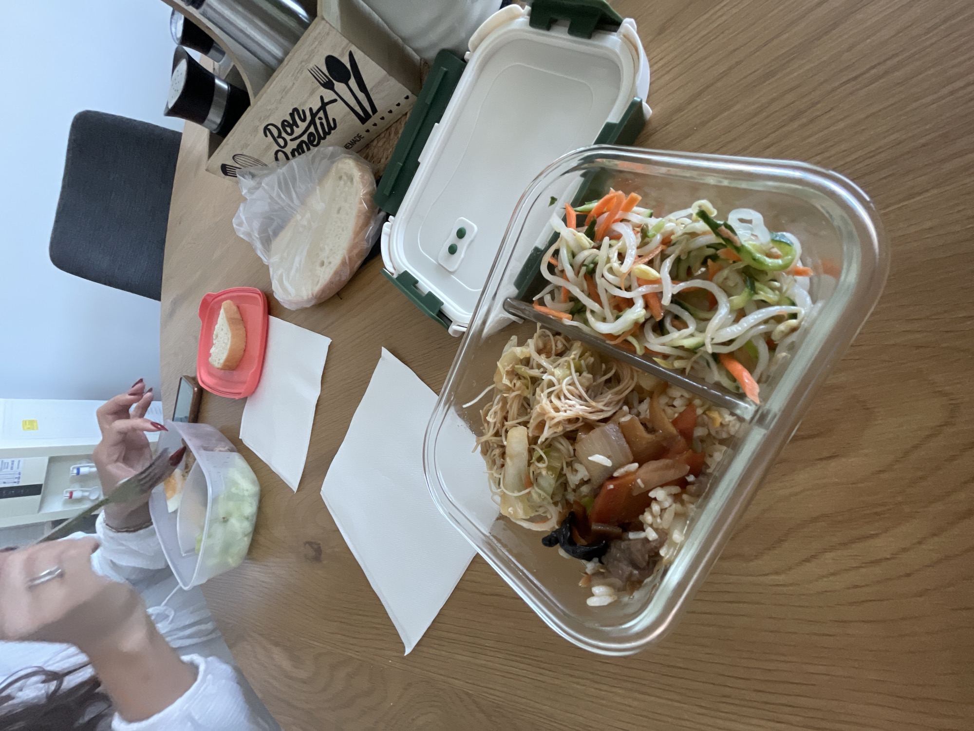 The image shows a wooden table setting with several items. A glass food container with two compartments holds different dishes: the top compartment contains julienned carrots, bean sprouts, and sliced cucumbers, while the bottom contains rice, vegetables, and meat. Next to the container is a white and green plastic lunchbox with the lid open. To the left of the lunchbox is a bread bag and a small container with a slice of bread inside. At the edge of the table, a woman is seen eating from a plastic container using a fork. The location is possibly in Sofia, Bulgaria. Other objects on the table include a container with the phrase "Bon Appetit" written on it, holding various items. The environment appears to be indoors, possibly a dining area.