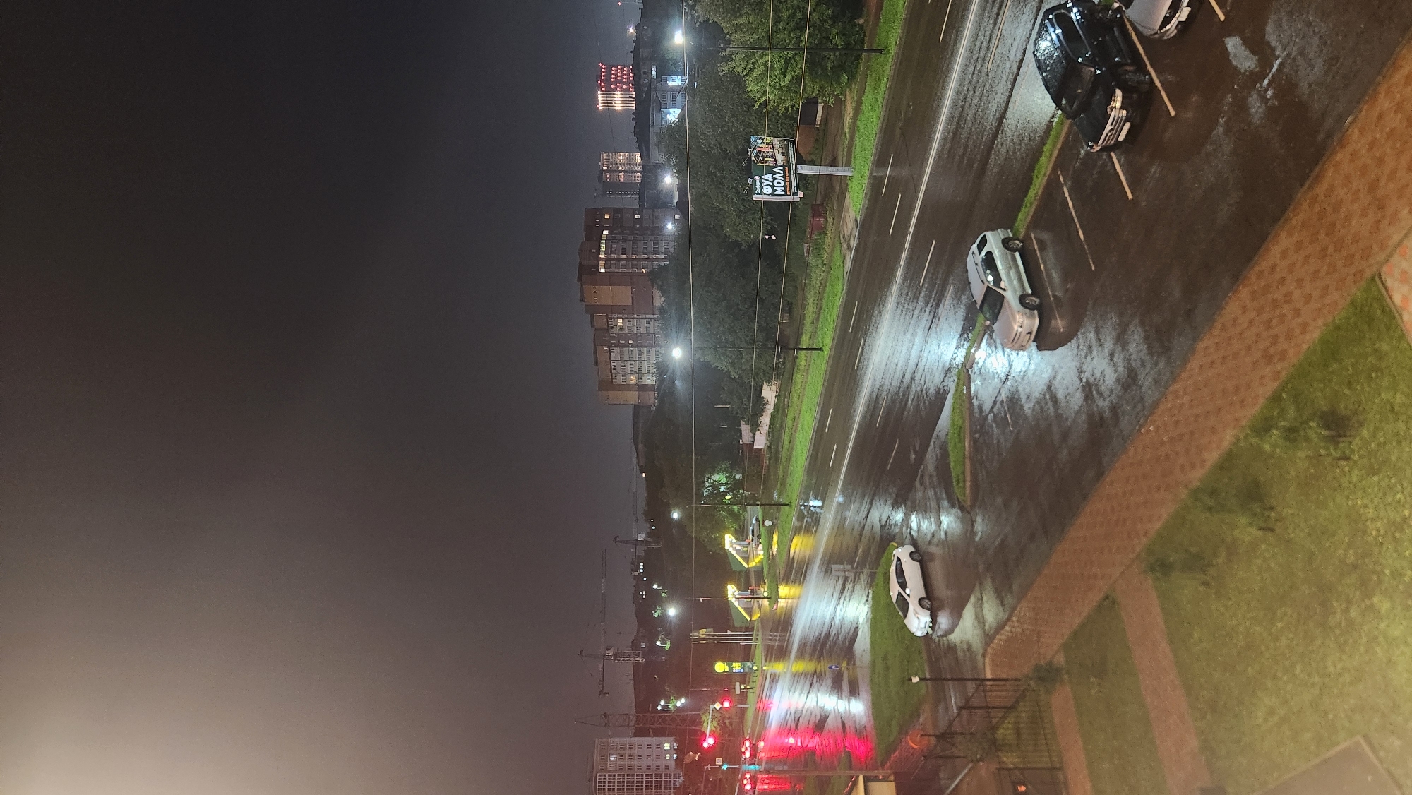 The image depicts a nighttime scene in an urban environment, likely during or shortly after a rainfall. A wet, multi-lane road dominates the foreground and midground, reflecting the lights of streetlamps and traffic signals. Several cars are visible on the road and in a parking lot on the right side of the frame. These include a white car in the left lane, a silver SUV in the parking lot, and a black SUV parked in a designated spot. A red traffic light is visible on the left side of the road.
In the background, apartment buildings and other structures are illuminated, some with lights on in their windows. Construction cranes are also visible in the distance. A billboard with text in Russian ("ФУД МОЛЛ") is positioned to the right of the road. The sky is dark and overcast, contributing to the overall dim lighting of the scene. The ground is wet, with puddles and reflections indicating recent precipitation. The composition is taken from an elevated perspective, looking down on the street and surrounding area.