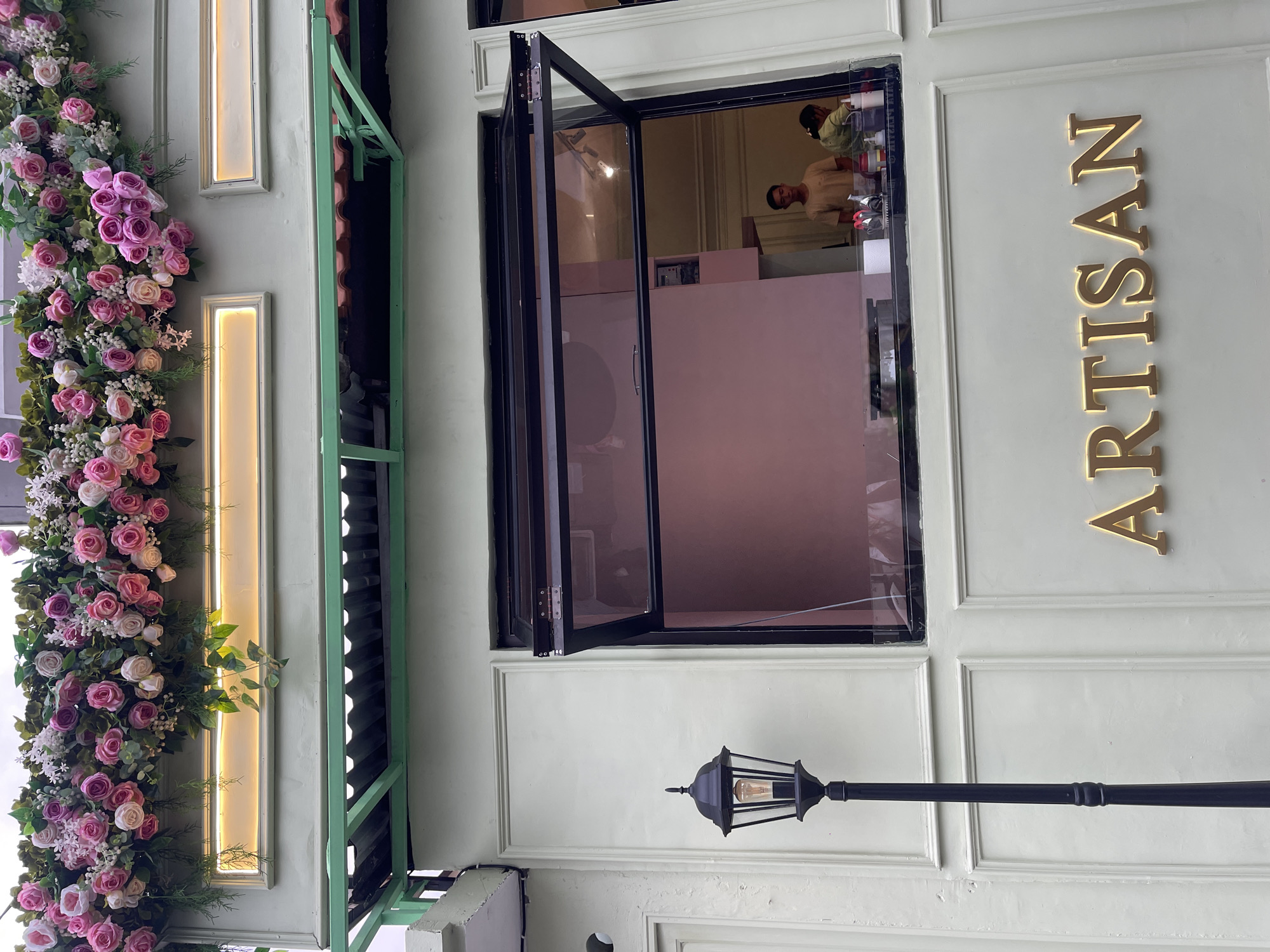 An exterior shot of a building in North Kuta, Indonesia, features a pastel green facade adorned with an elaborate floral arrangement of pink and white roses and greenery at the top. Below this, a rectangular neon light illuminates a section of the facade. Further down, a dark green metal awning extends over a window. The main focus is a large, open window with a black frame, revealing the interior of a shop. Through the window, two people are visible behind a counter; one man in a beige t-shirt stands and looks forward, while a woman in a green shirt is partially visible, attending to something. The shop's interior wall is a soft pink. To the left of the window, a black vintage-style lamppost stands tall against the wall. Below the window, large, gold-colored, illuminated letters spell out "ARTISAN" on the pastel green wall. The building's facade has decorative paneling.