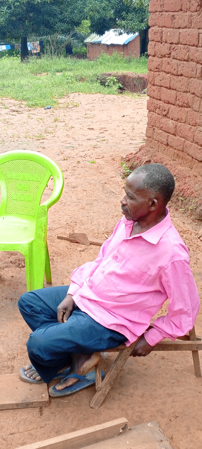 An outdoor, daylight scene in Ngalula, Democratic Republic of the Congo, features a single adult male seated on a low wooden stool, positioned in the lower-right quadrant of the frame. He is dark-skinned, with short, dark hair and a mustache, and wears a pink long-sleeved button-up shirt with sleeves rolled to the forearms, dark blue trousers, and blue flip-flops. His body is oriented towards the right, and his head is turned slightly to the right, looking off-frame. His left hand rests on the wooden stool.
To the left of the man, a bright green plastic monobloc chair with the text "GRACE OF GOD" embossed on its backrest stands empty. The ground is reddish-brown dirt or earth, with sparse patches of green vegetation. In the immediate foreground on the right, a wall constructed of reddish-brown bricks is visible.
In the mid-ground and background, dense green foliage from trees and shrubs is present. Underneath a large tree in the background, a clothesline displays colorful garments hanging to dry. Two small, single-story buildings with reddish-brown walls and light-colored roofs featuring blue trim are visible further in the background, partially obscured by vegetation. Scattered wooden debris lies on the dirt ground near the man's feet and behind the green chair.