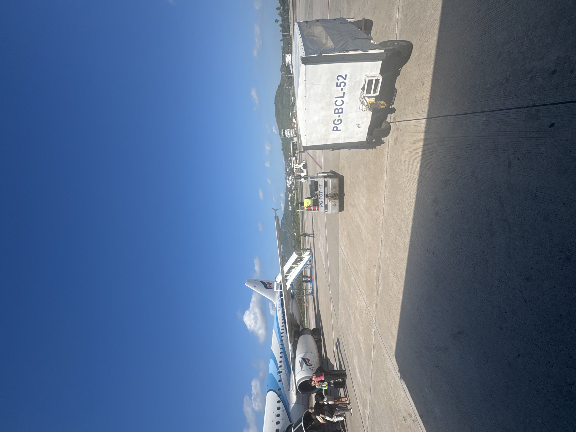 The image depicts an airplane parked at an airport in the city of บ้านบางรักษ์, Thailand. The scene includes an aircraft, likely a commercial passenger plane, with a boarding ramp attached. People are visible near the entrance of the aircraft, potentially boarding or deboarding. Additionally, there are cargo or baggage handling vehicles and equipment on the tarmac, one labeled "PG-BCL-52". The sky is clear and blue, and the background shows vegetation and possibly a small mountain. The image is taken from ground level, possibly from a passenger's perspective.