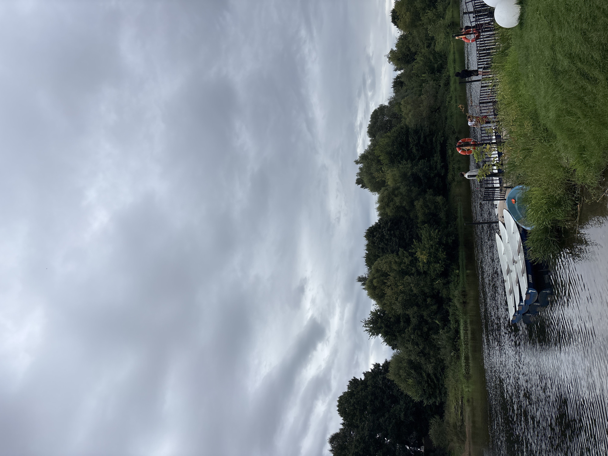 A wide shot shows a cloudy sky over a tranquil river and a lush green riverbank in Zvenigorod, Russia. Several white boats are tied to a wooden dock, where four people are standing and observing the water. The figures are dressed casually, and some wear life vests. The river is calm, with gentle ripples on its surface, and it reflects the overcast sky. Dense green trees line the opposite bank of the river, creating a natural backdrop to the scene. The foreground features a grassy bank dotted with wildflowers. The overall atmosphere is serene and natural.